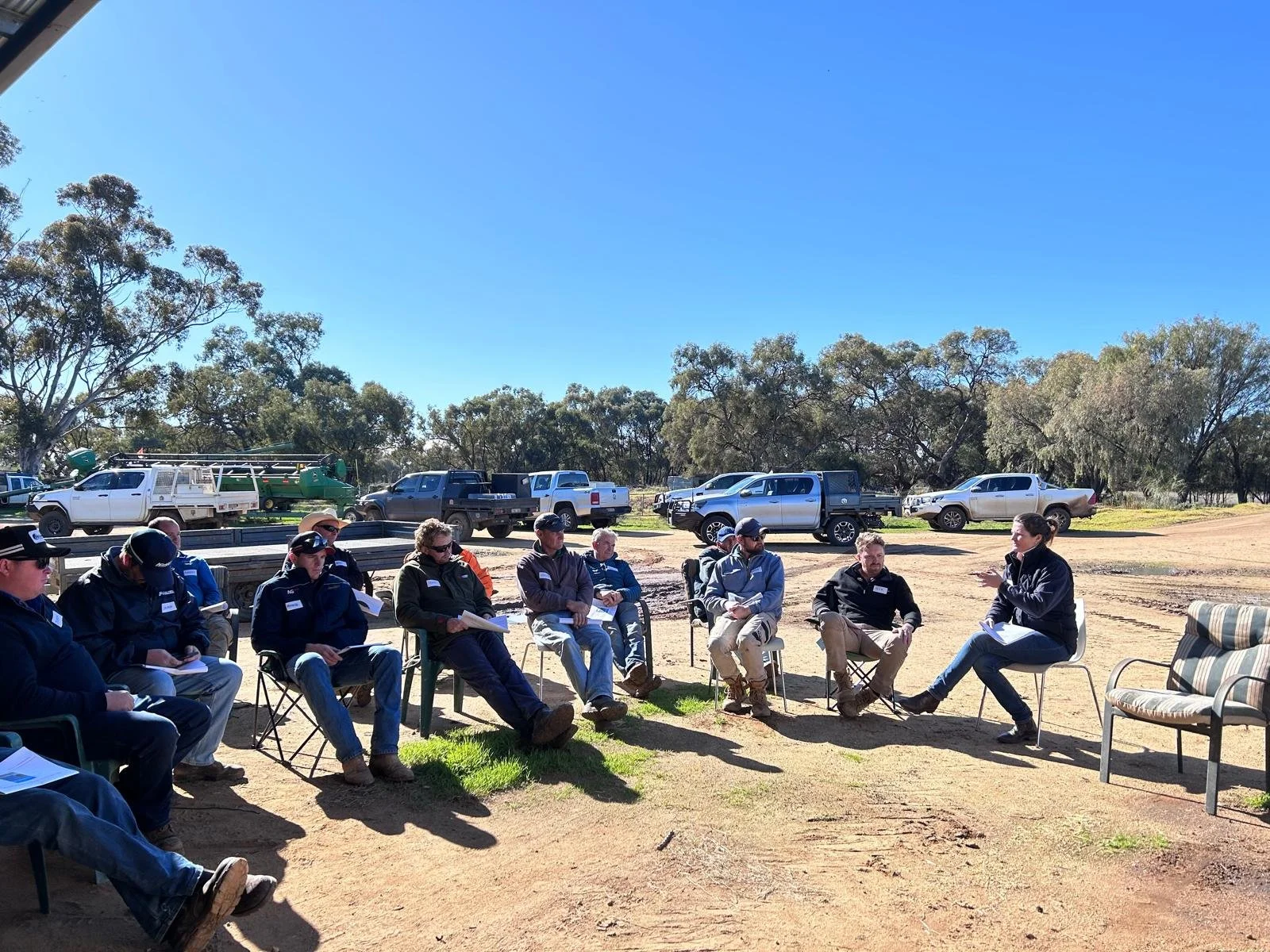 Group of people in outdoor meeting, sitting on chairs in a semi-circle, with parked trucks and trees in the background on a sunny day.