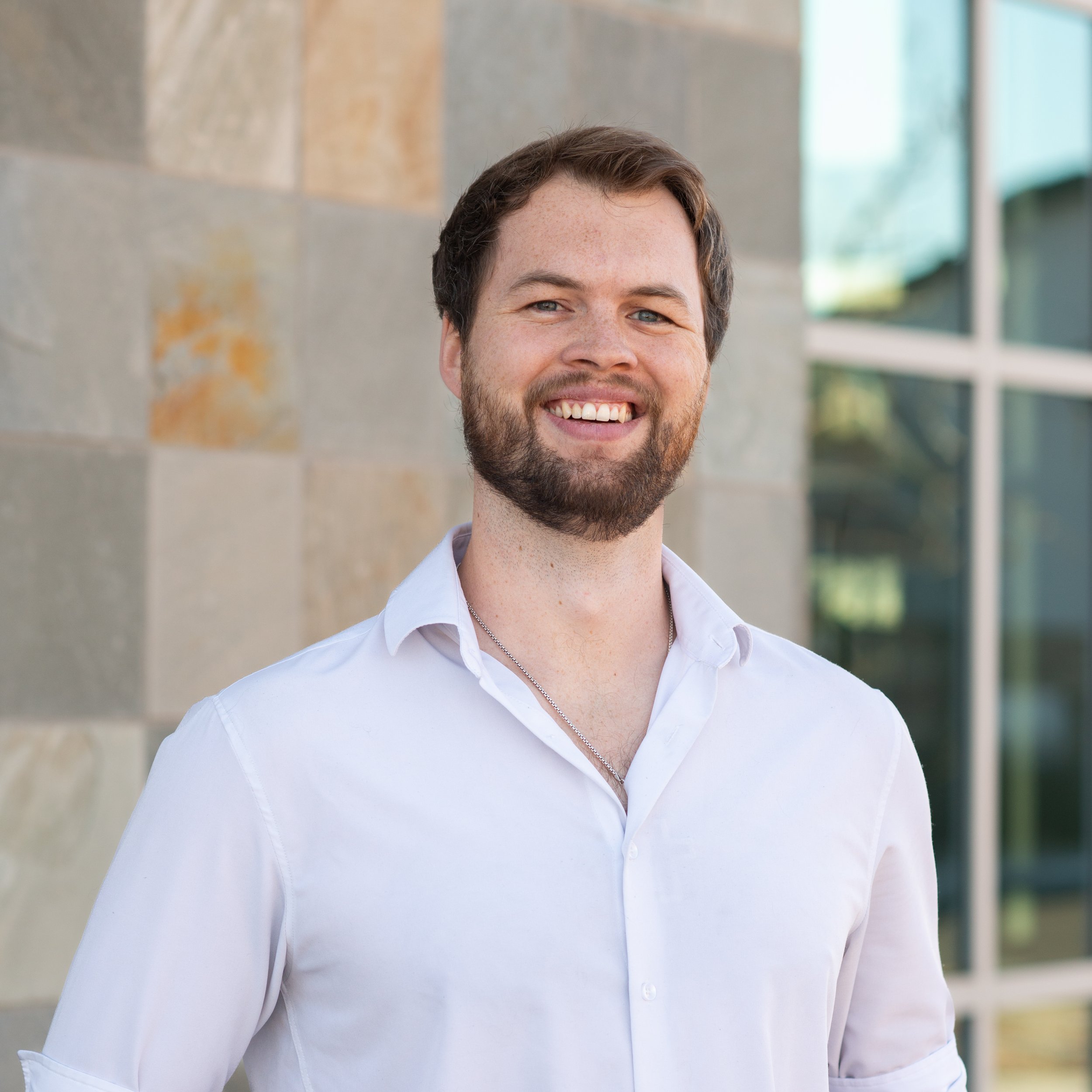 A young man with dark hair and a beard, wearing a white button-down shirt, smiling outdoors in front of a building with large windows and stone walls.