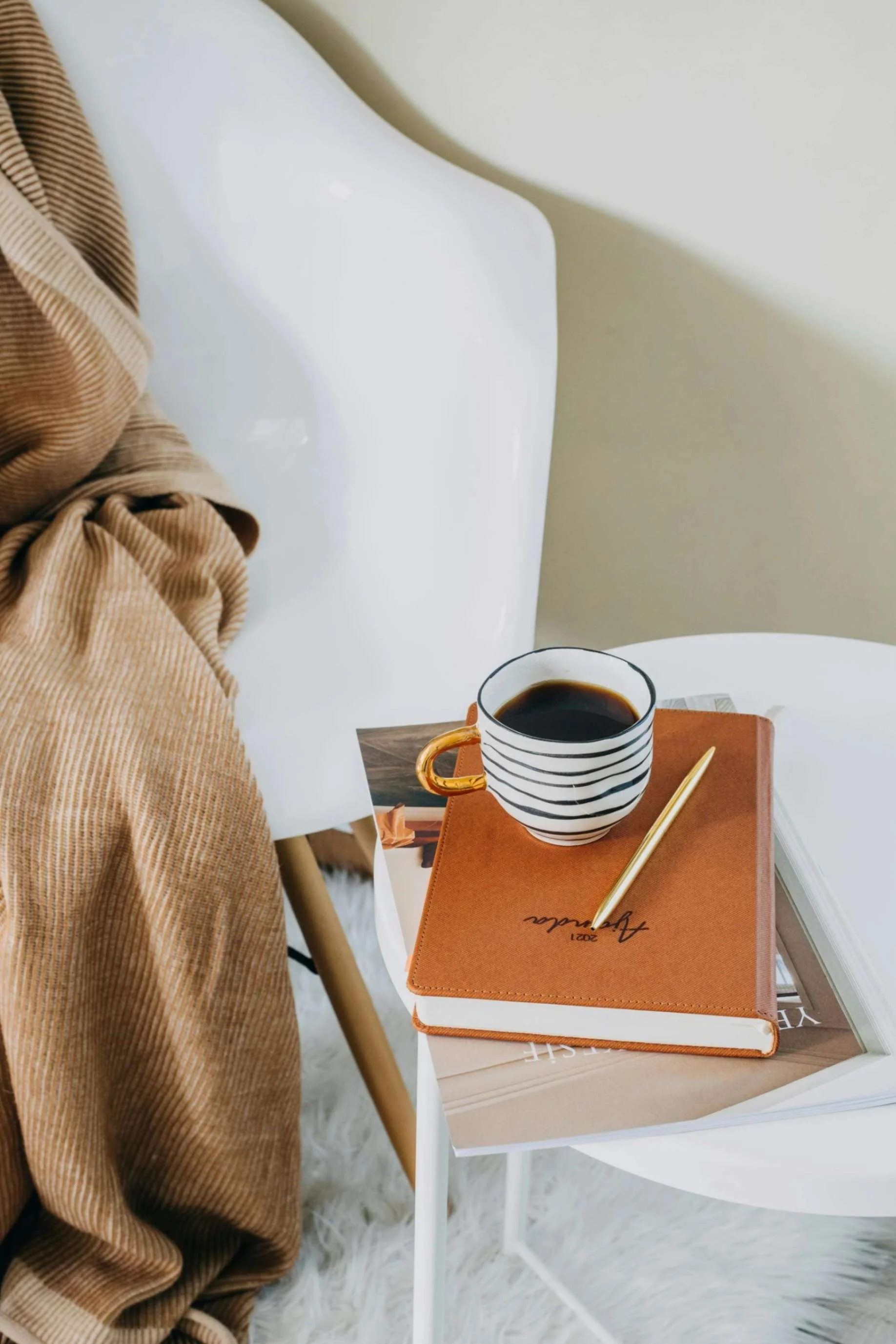 A white bedside table with a striped coffee mug, a gold pen, and a brown leather journal resting on top. Part of a white chair and a person wearing a brown corduroy jacket are visible in the background.