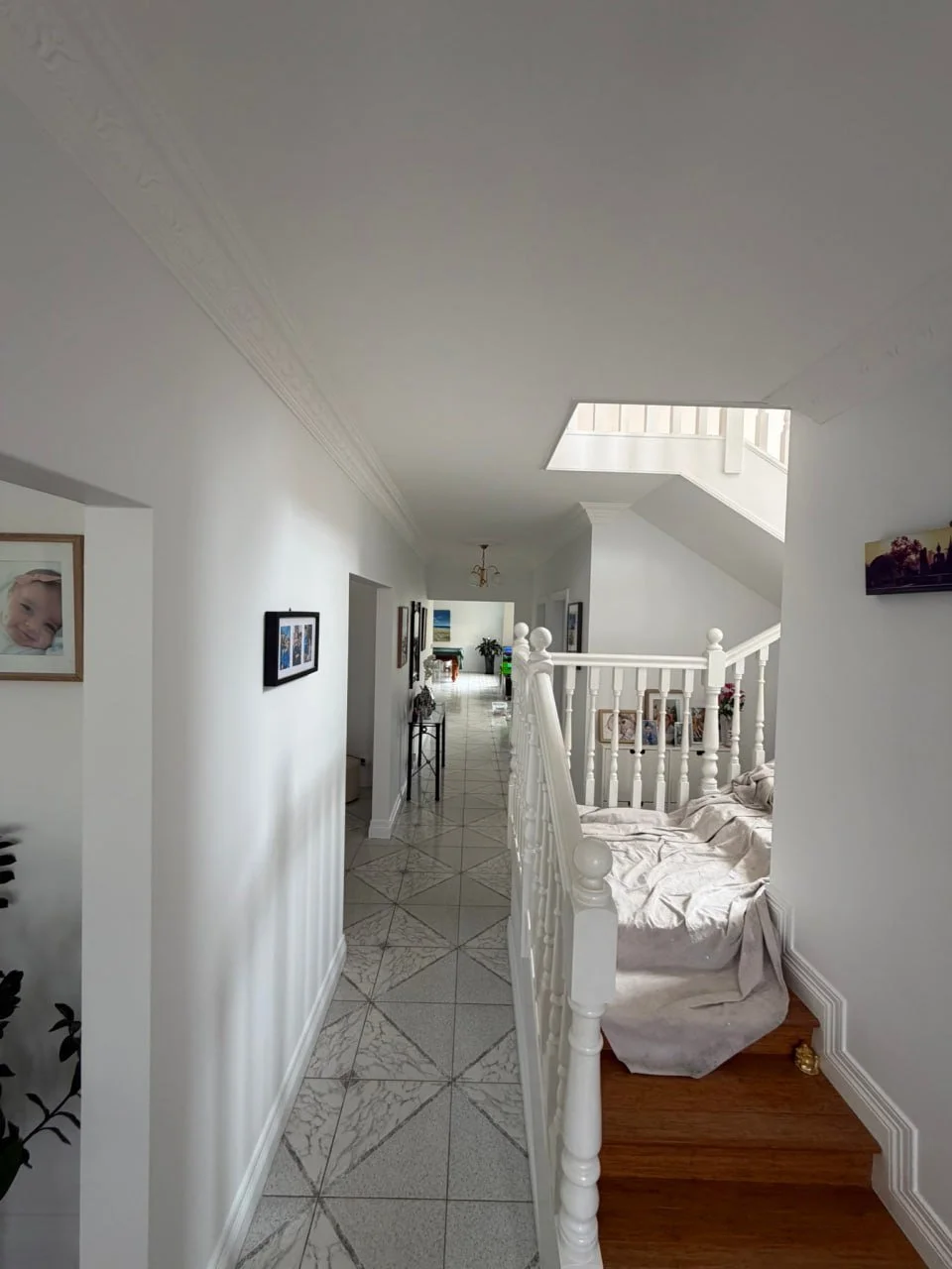 Interior view of a bright, white corridor in a house with framed artwork on the walls, a staircase leading to upper floor, and a couch with a beige cover.