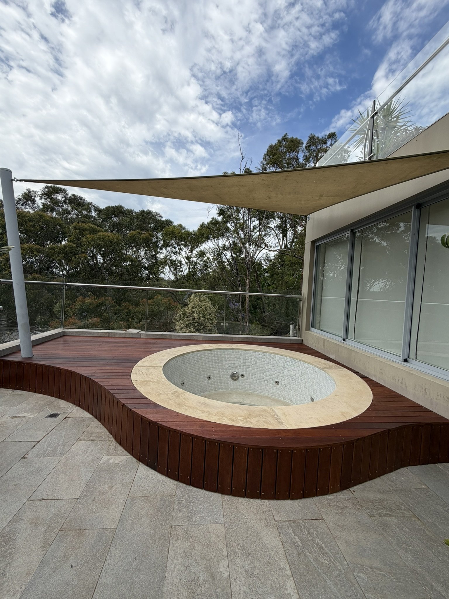Outdoor balcony with a circular hot tub set into a wooden deck, surrounded by a glass railing, with view of trees and a partly cloudy sky.