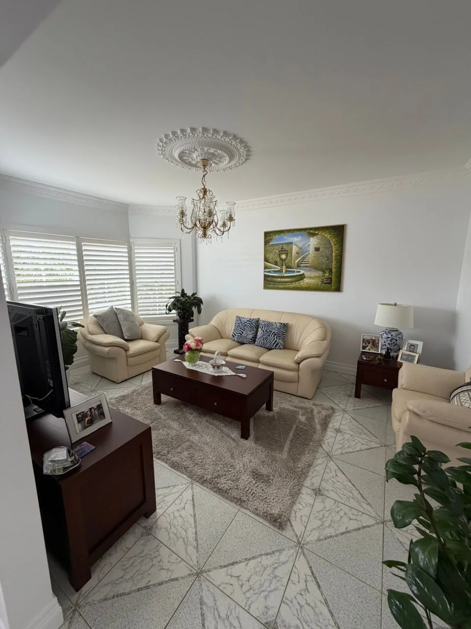 Bright living room with white walls, cream-colored sofas, a wooden coffee table, a TV stand, a chandelier, and framed family photos. There are large windows with white blinds, a painting on the wall, and potted plants.