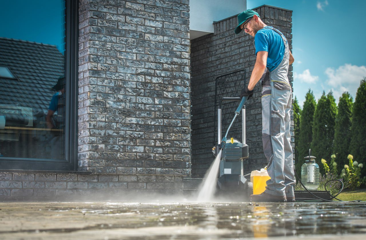 Man pressure washing a wooden deck outside a house with brick walls and large windows on a sunny day.