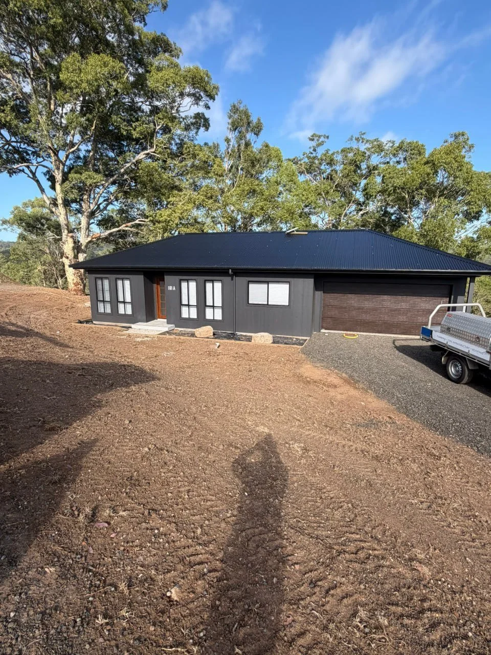 Newly built single-story house with black exterior, metal roof, and attached garage, surrounded by a dirt yard and trees in the background, under a partly cloudy sky.