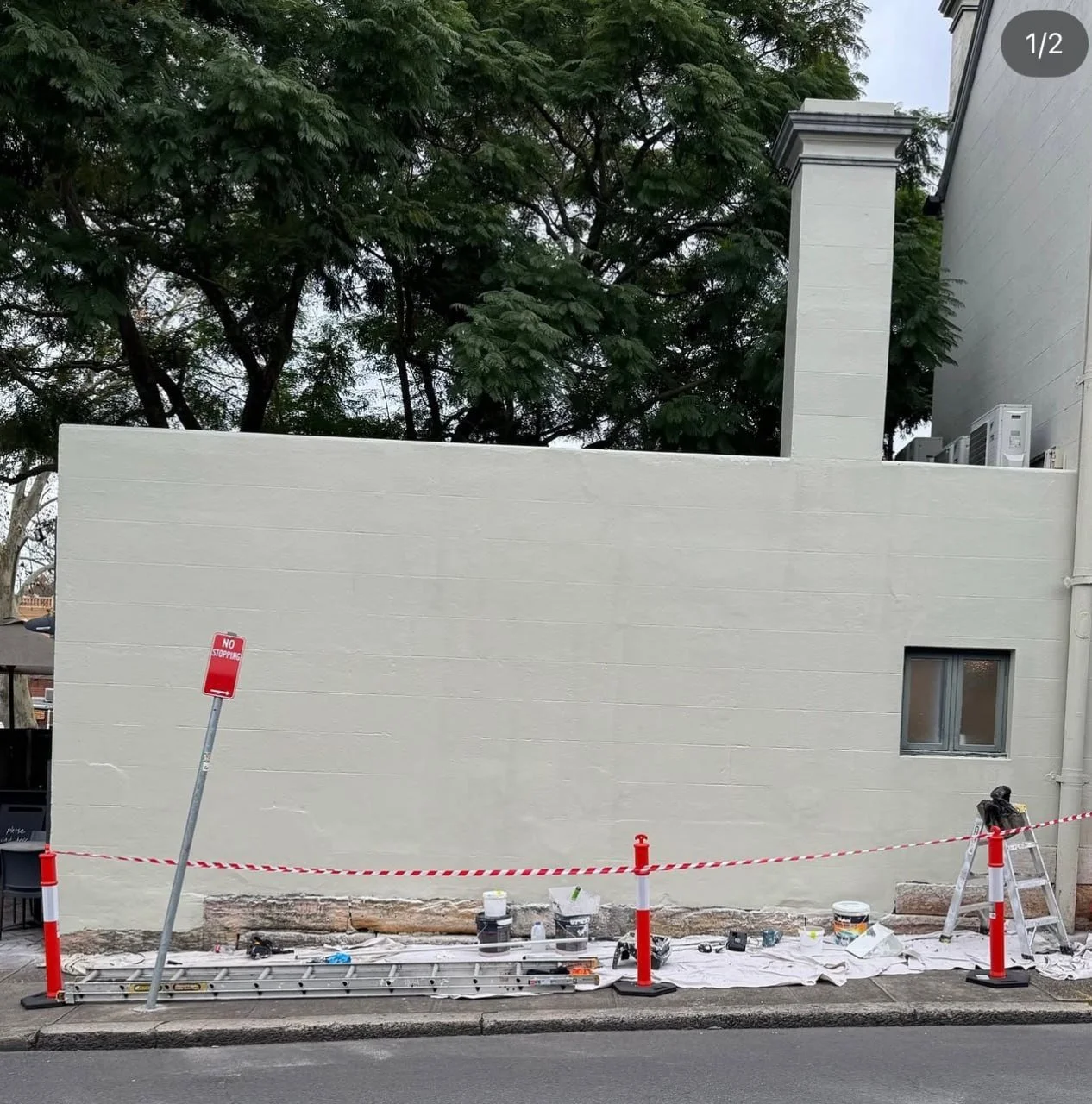A newly painted white wall in front of a sidewalk, with construction tools, a ladder, paint cans, and a 'No Stopping' street sign, all surrounded by orange and white barrier tape.