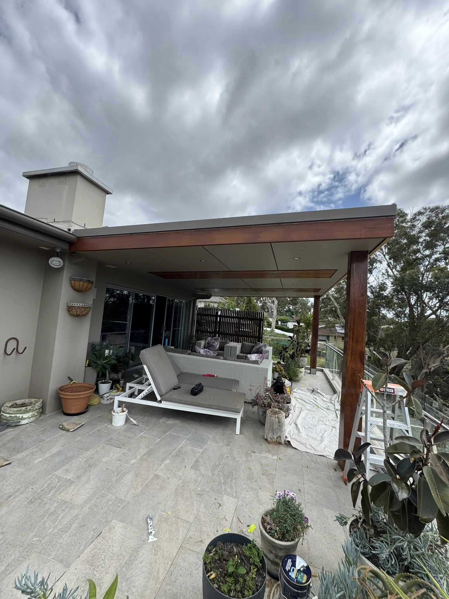 An outdoor patio area under renovation with furniture, potted plants, a ladder, and construction materials, with a cloudy sky overhead.