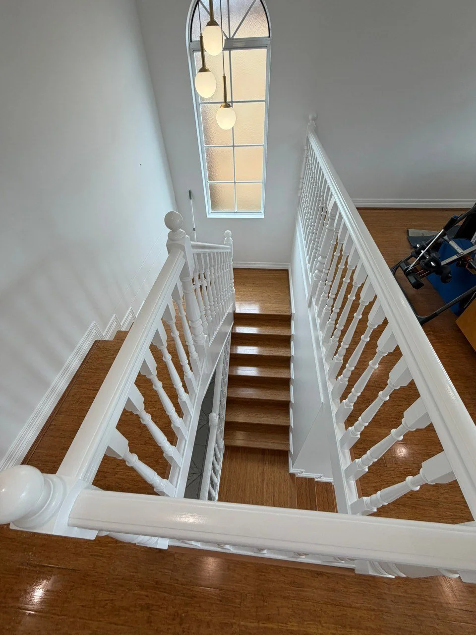 Interior view of a wooden staircase with white balustrades, seen from the top looking down. There is a tall, arched window at the landing, and a hanging light fixture with three bulbs. Part of a room with hardwood floors and some furniture or equipment is visible.