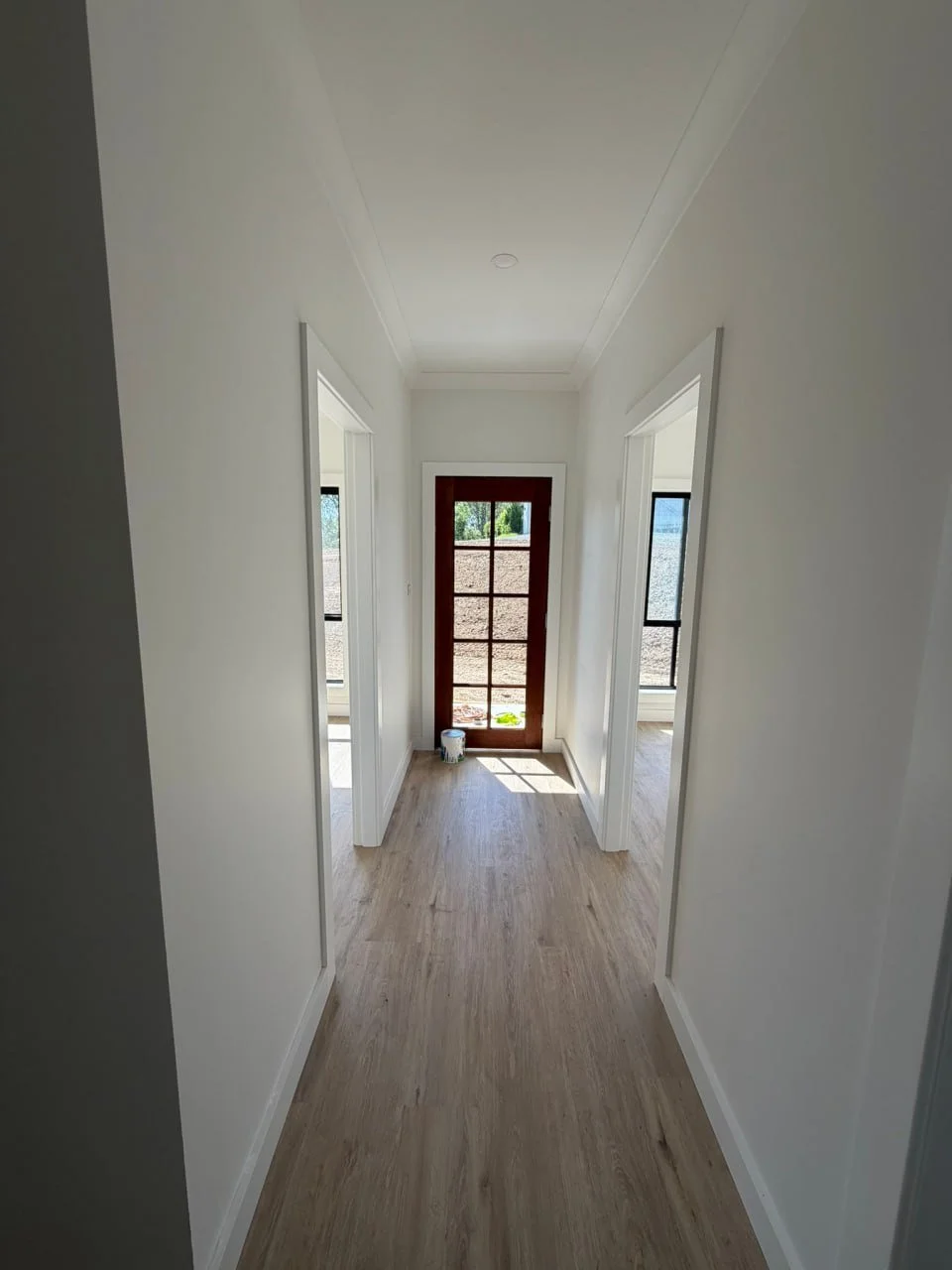 Empty hallway with white walls, wooden flooring, and natural light from windows and a door at the end.
