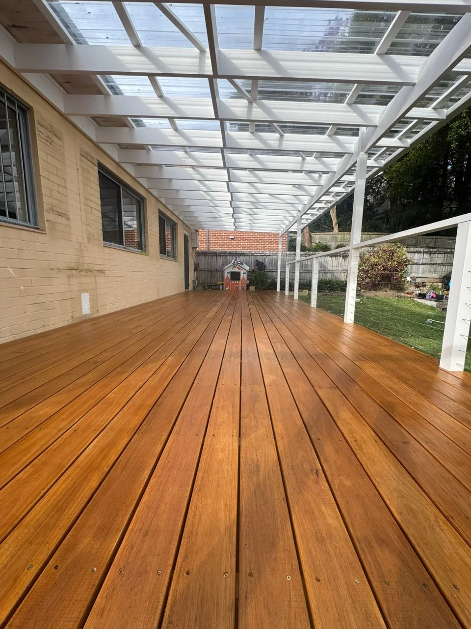 A spacious outdoor wooden deck with a transparent roof overhead, attached to a brick house with windows. The deck is bordered by a white railing on the right side and leads to a small playhouse in the backyard.