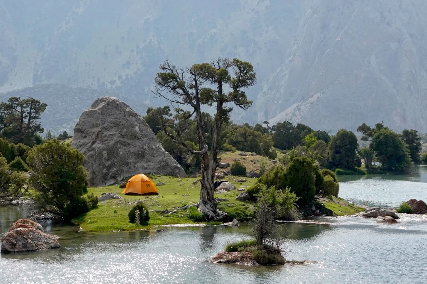 A lakeside scene with a small orange tent pitched on green grass near a large rock and a twisted tree. The lake has small rocks and bushes along the shore, with a mountain range in the background.