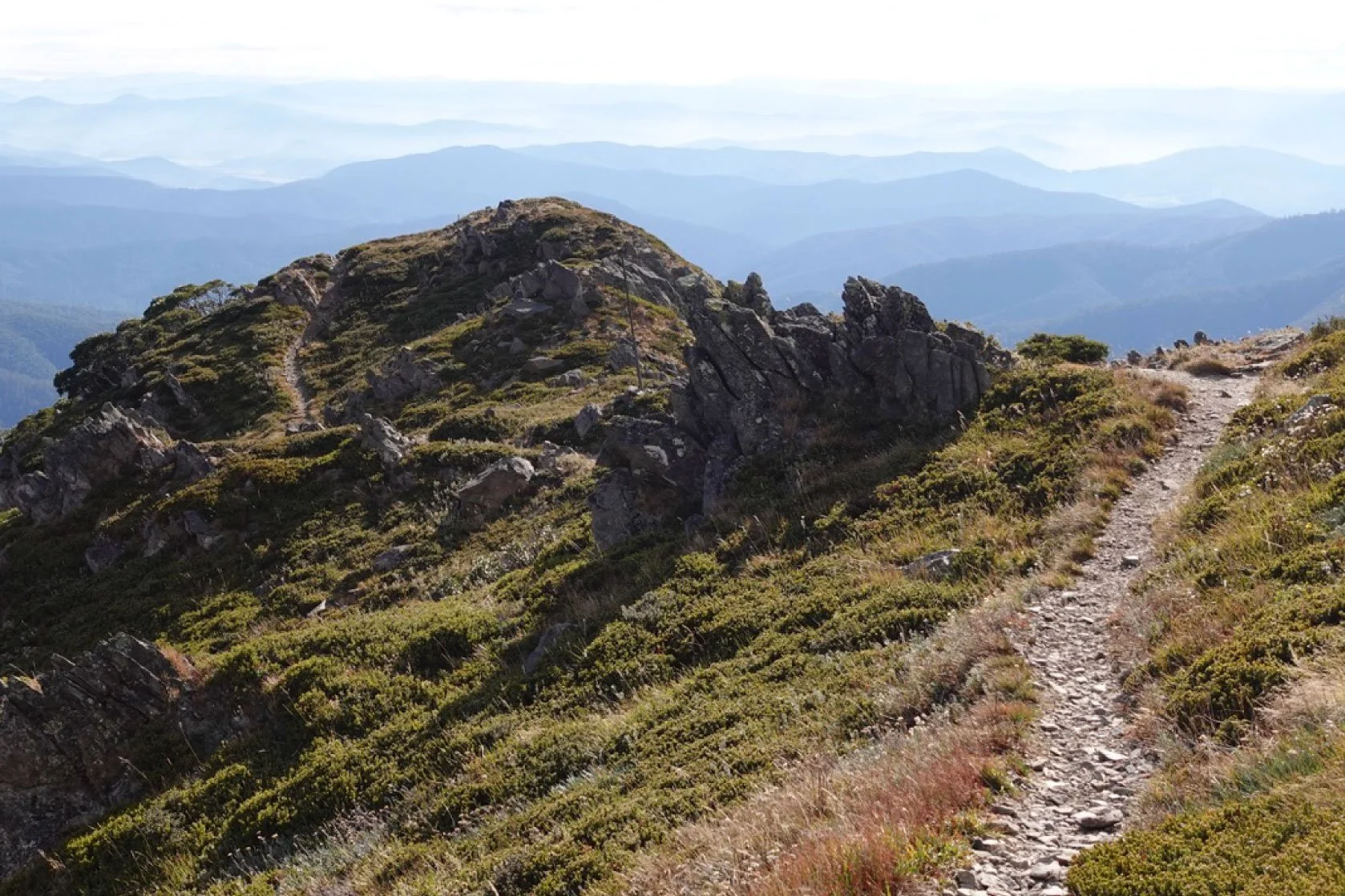 A mountainous trail with rocky terrain and lush green vegetation leading up to a summit with layered mountain ranges in the background.