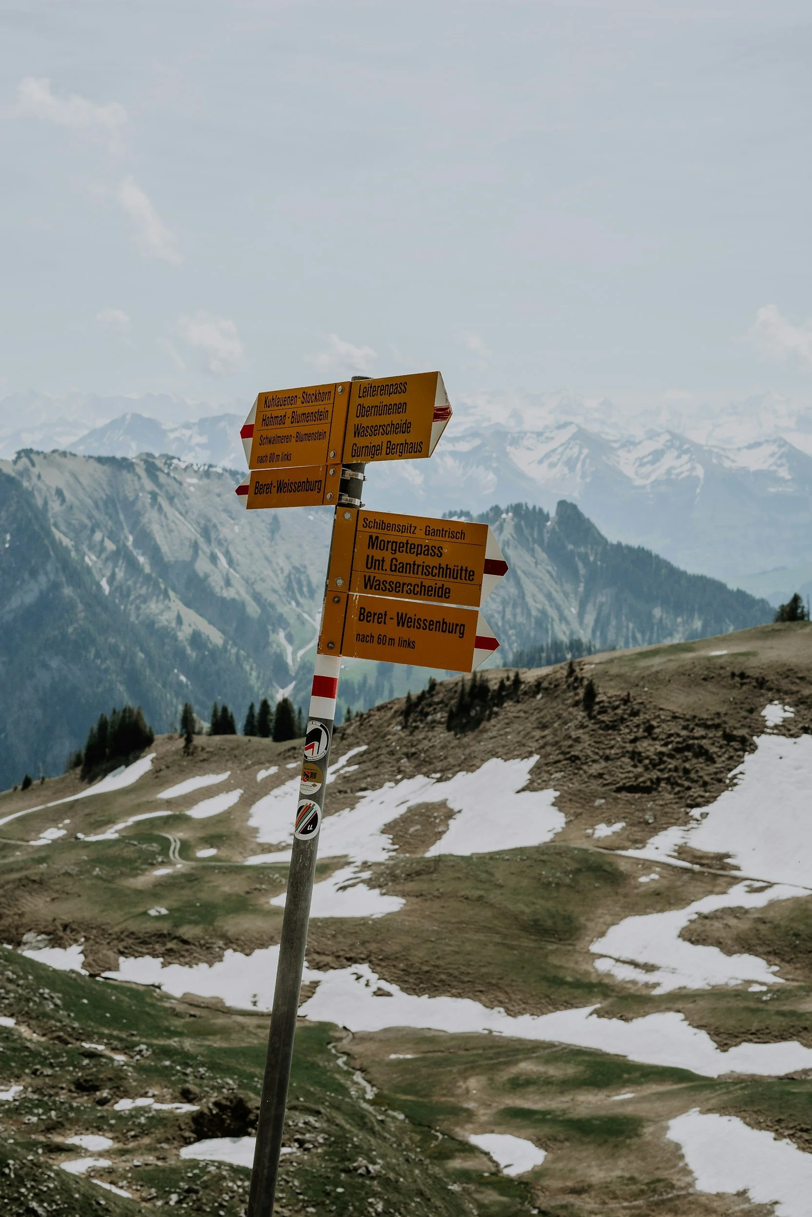 A colorful directional signpost stands in a mountainous landscape with snow patches, green valleys, and distant snow-capped peaks under partly cloudy skies.