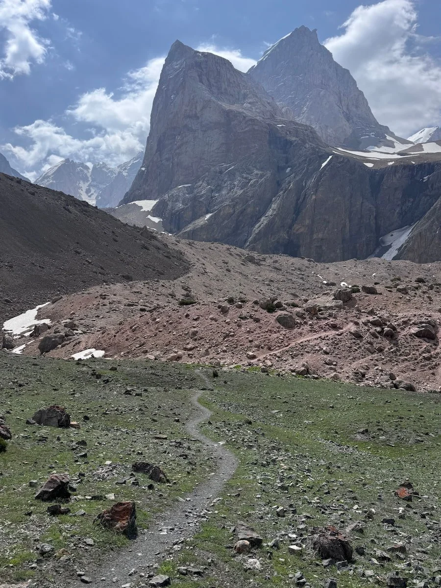 A mountainous landscape with a large, rocky peak in the background, partly snow-capped, and a small, winding dirt trail in the green foreground.