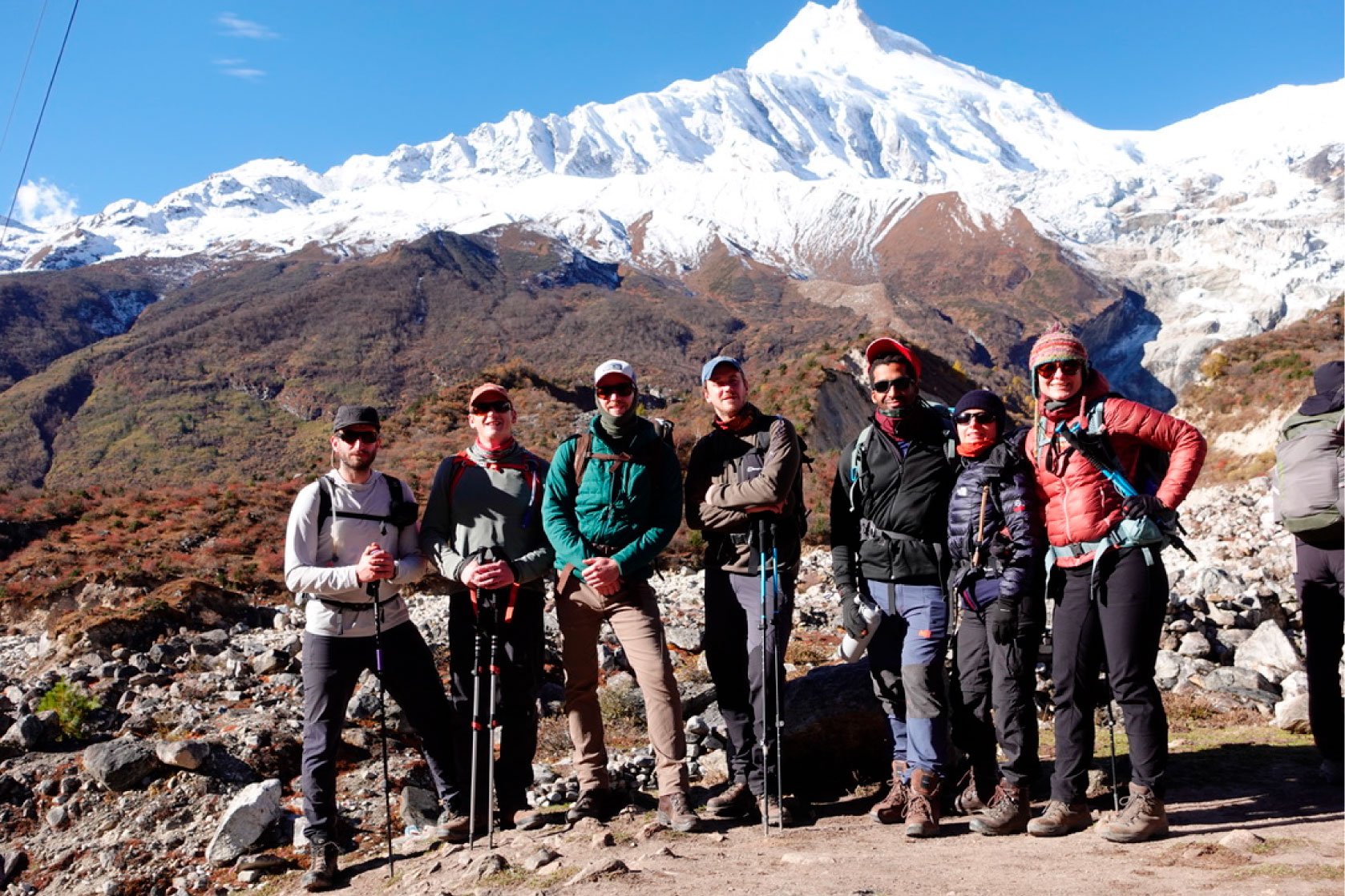 A group of eight hikers standing on a rocky trail with a snow-capped mountain in the background. The hikers are dressed in outdoor gear, including jackets, hats, sunglasses, and backpacks, and are holding trekking poles. The mountain behind them is tall with a sharp peak and snow-covered slopes under a clear blue sky.