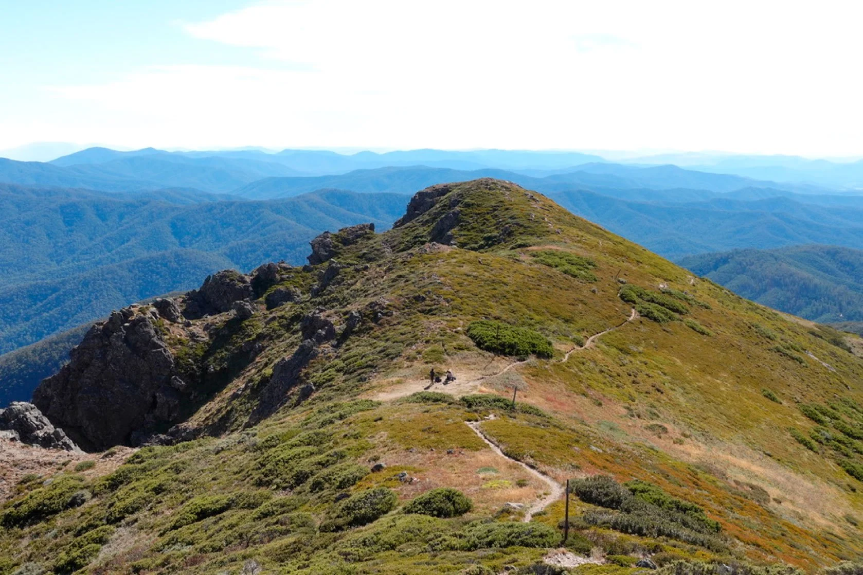 A trail winds along a mountain ridge with hikers resting near the path, surrounded by green and brown shrubbery, with distant mountain ranges under a partly cloudy sky.