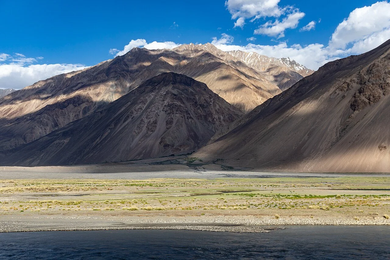 Mountain landscape with rugged, barren peaks and a river in the foreground under a partly cloudy sky.