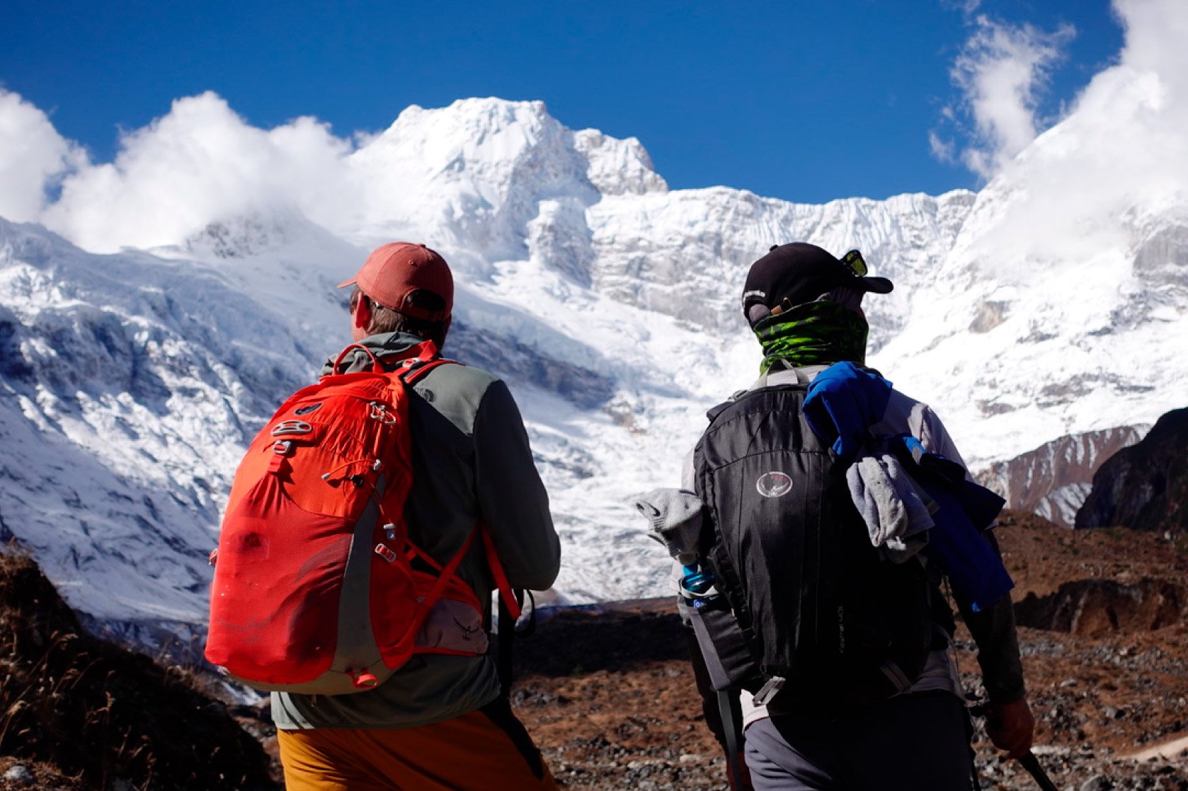 Two hikers with backpacks standing on a rocky terrain, looking at snow-covered mountains and glaciers under a blue sky.