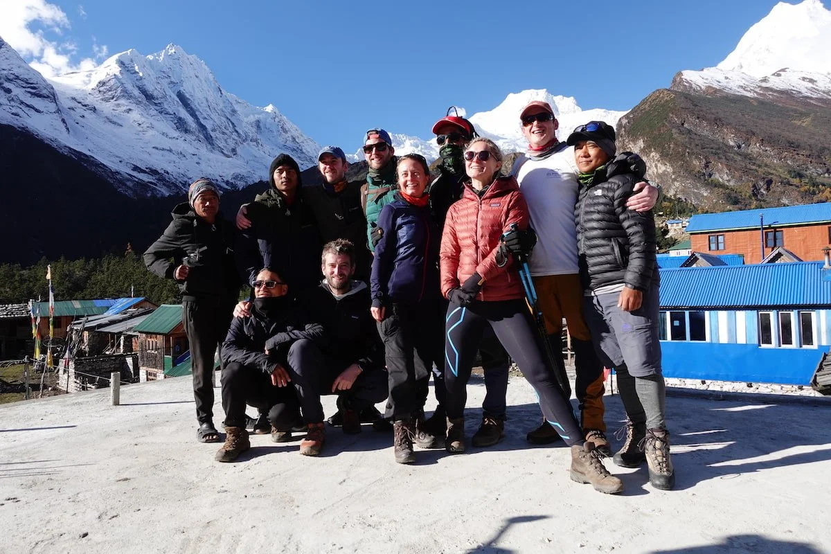 Group of people in outdoor winter gear posing together in a mountainous area with snow-capped peaks and blue sky in the background.