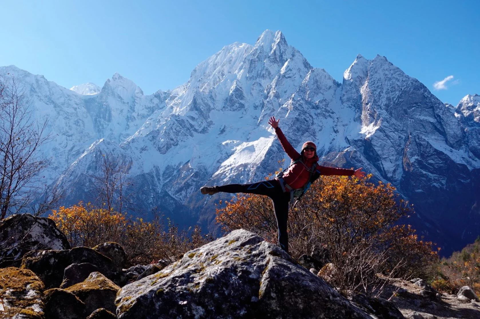 A woman in outdoor gear balancing on a rock with one leg extended sideways in front of snow-capped mountains under a clear blue sky.