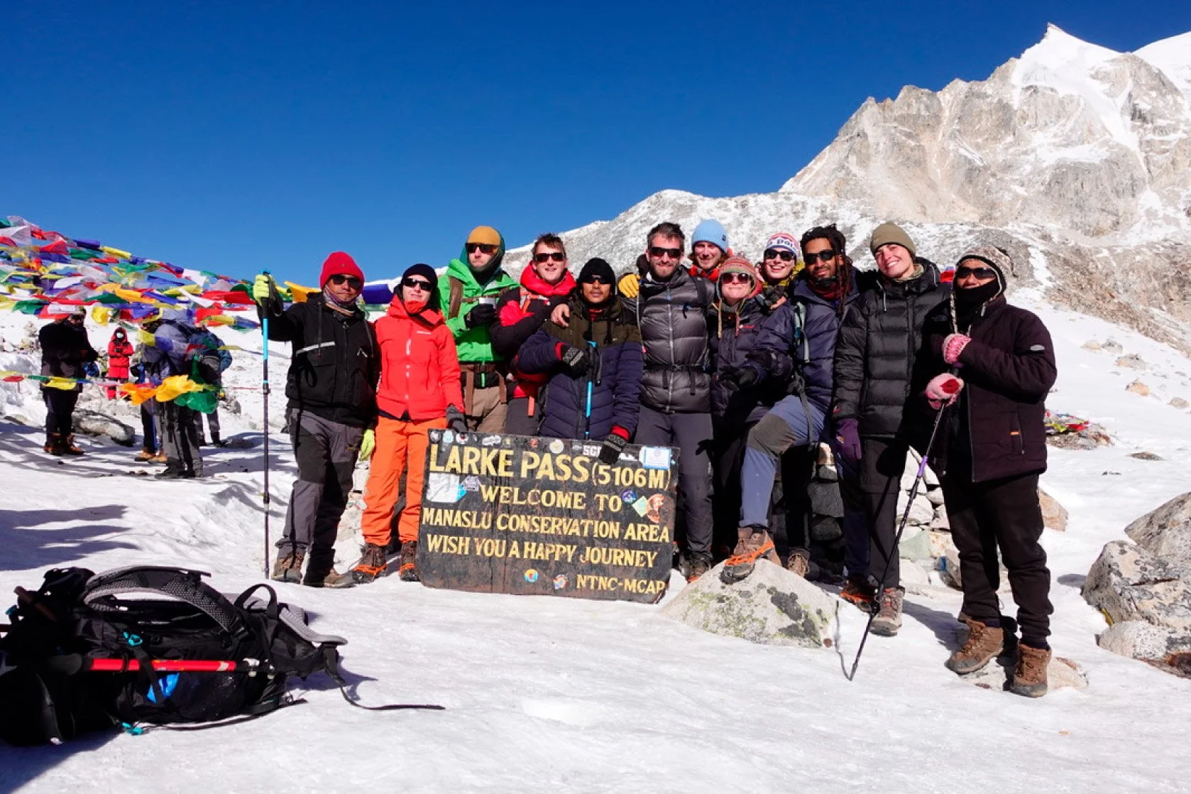 Group of climbers standing on snow at Larke Pass (5106 meters), holding a sign that welcomes visitors to Manaslu Conservation Area, with colorful prayer flags and snow-covered mountains in the background.