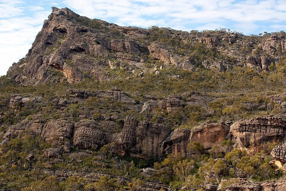 A rugged mountain landscape with layered rock formations and sparse vegetation under a partly cloudy sky.
