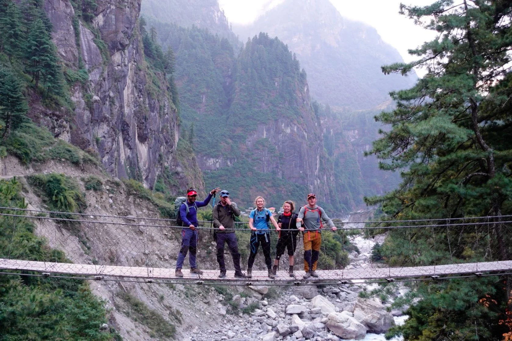 Group of five hikers standing on a suspension bridge in a mountainous forest landscape.