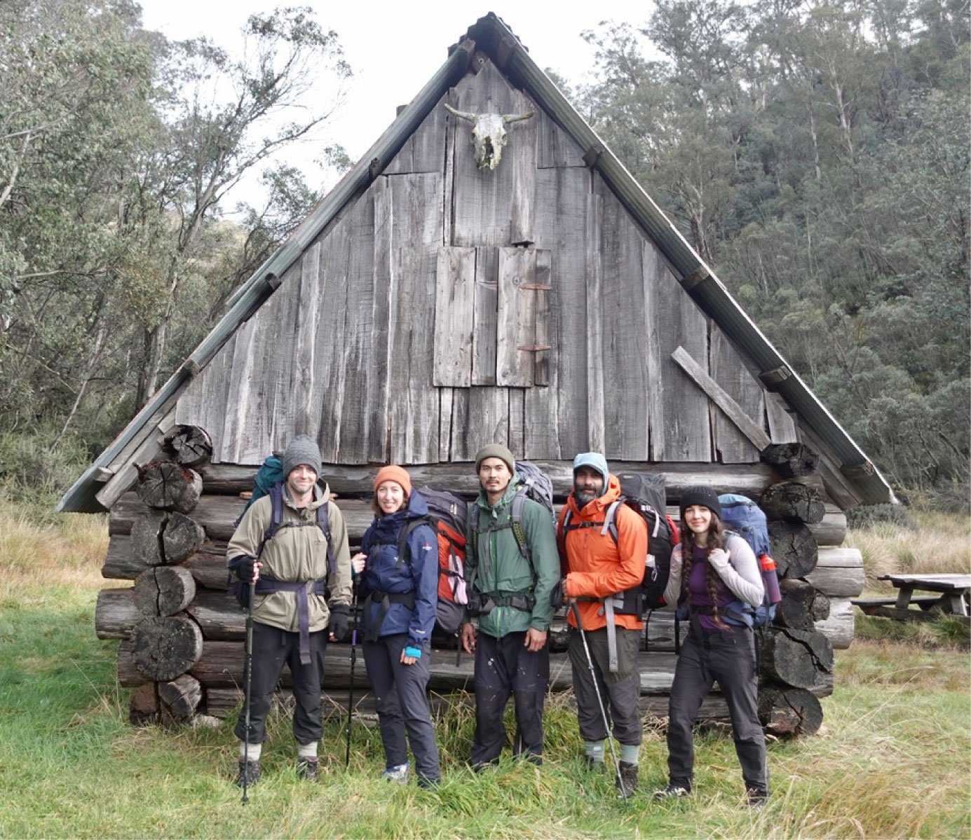 Group of five hikers standing in front of a rustic wooden cabin in a forested area.