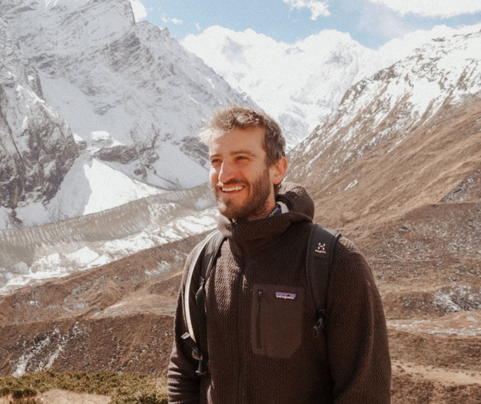 A man with a beard and short hair, dressed in outdoor hiking gear with a backpack, smiling in front of snowy mountains and a partly cloudy sky.