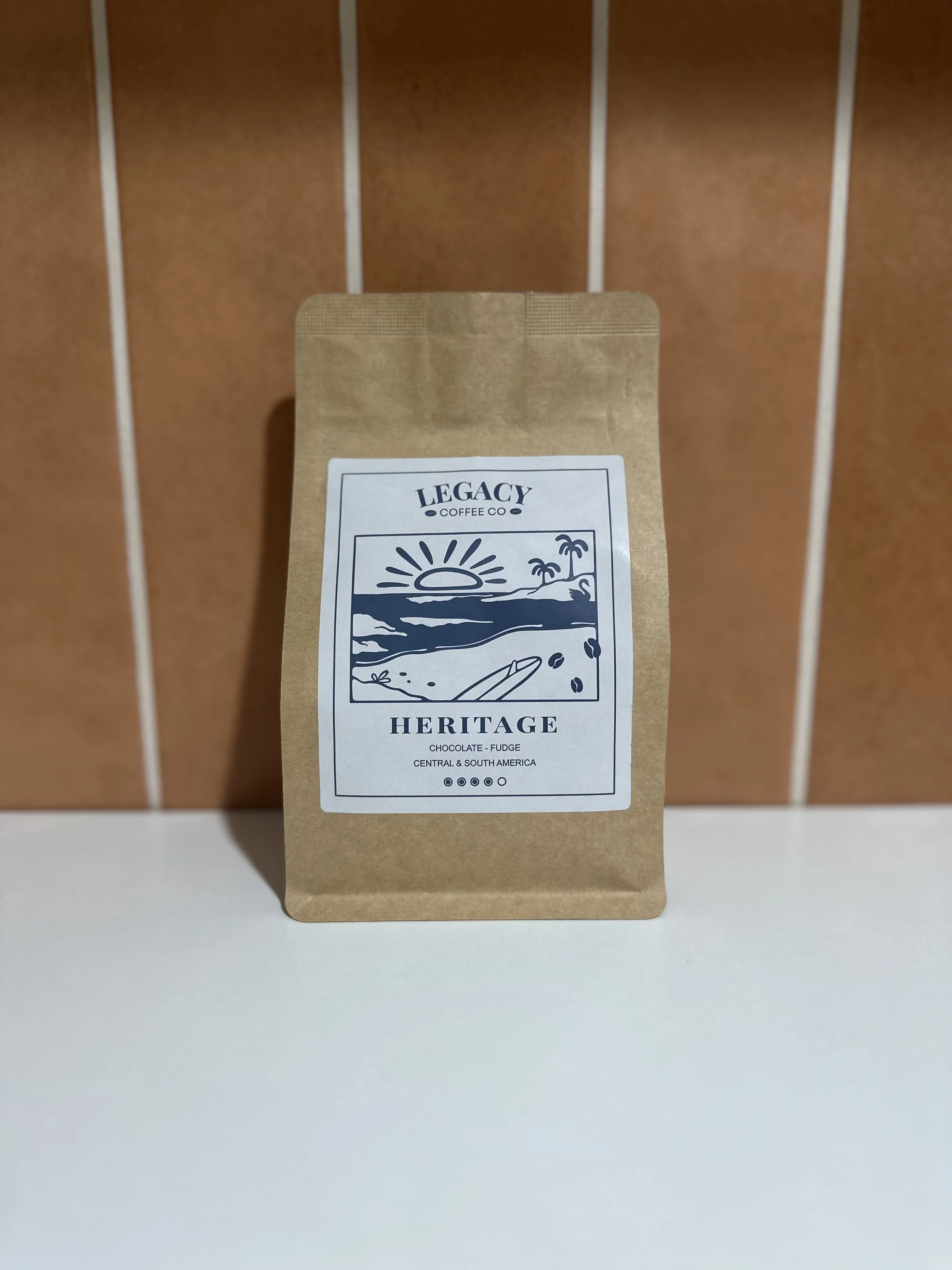 A brown paper bag with a white and blue label reading "Legacy Coffee Co. Heritage." The label features a drawing of a sunset over the ocean with palm trees, and mentions "Chocolate - Fudge" from "Central & South America."