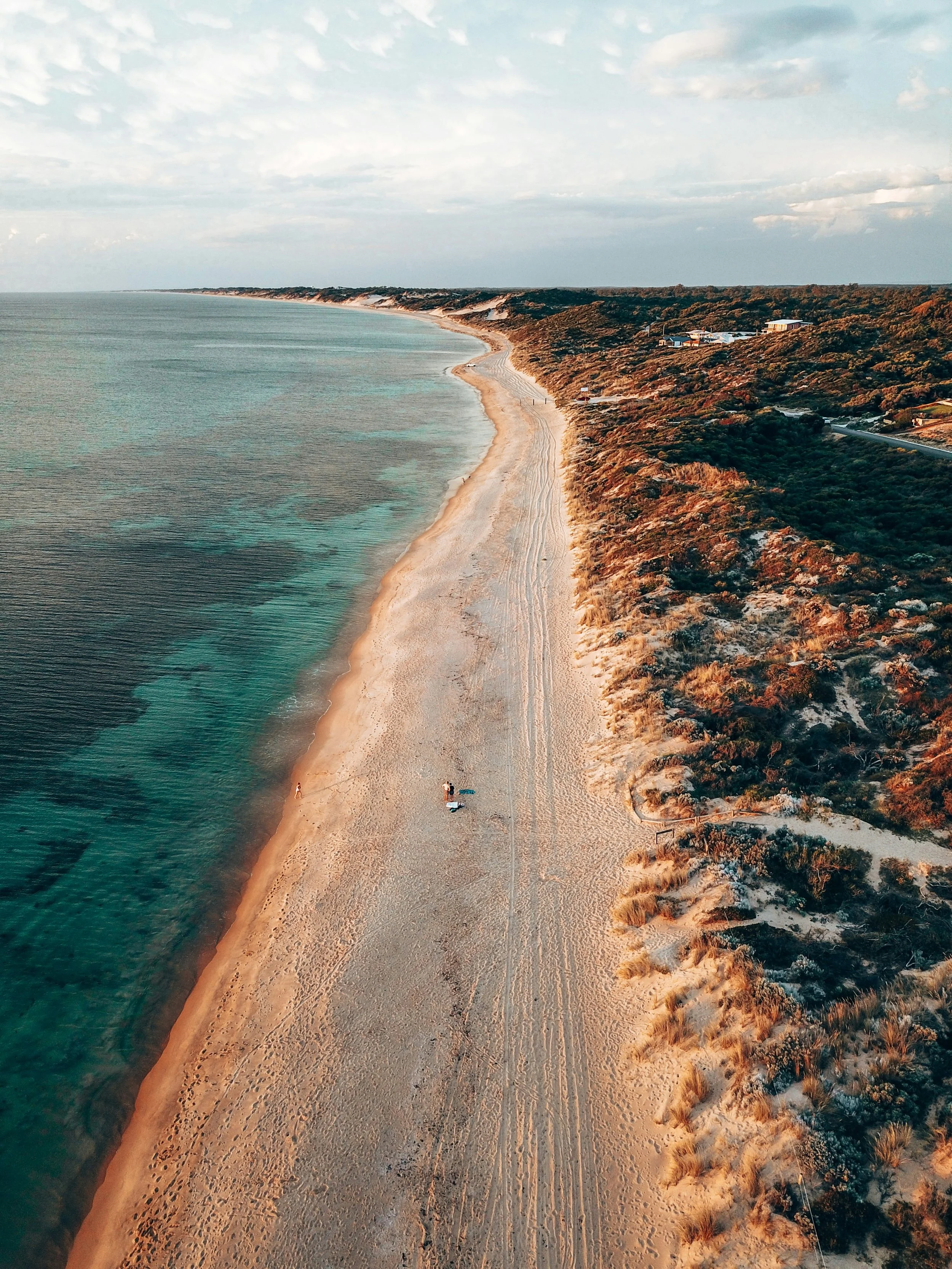 An aerial view of a long, curving beach with turquoise waters on one side and sandy dunes with sparse vegetation on the other, with a few people on the beach and some buildings on the dunes.