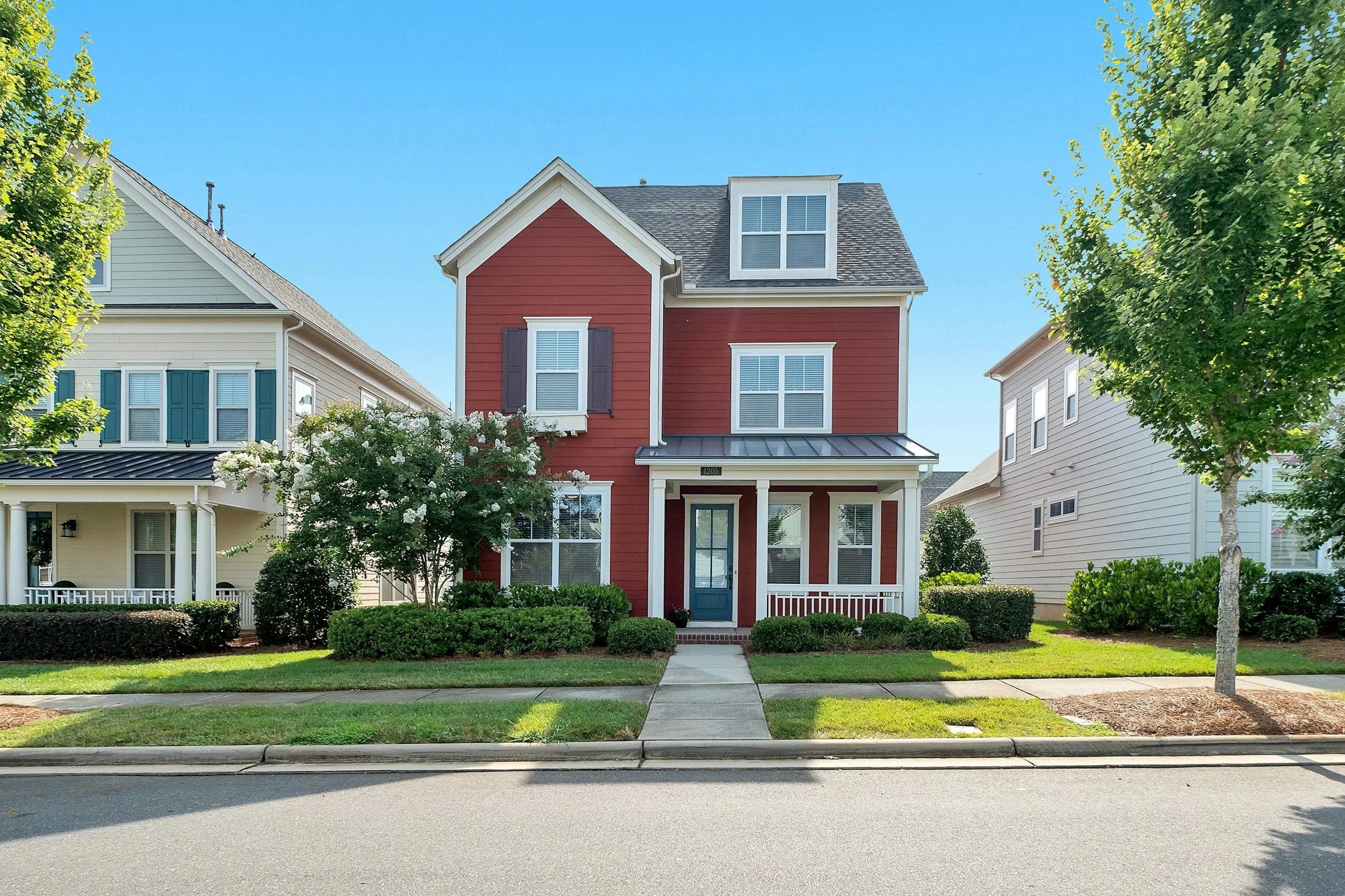 A three-story red house with white trim and a blue door, surrounded by green bushes and trees, located between two other homes under a clear blue sky.