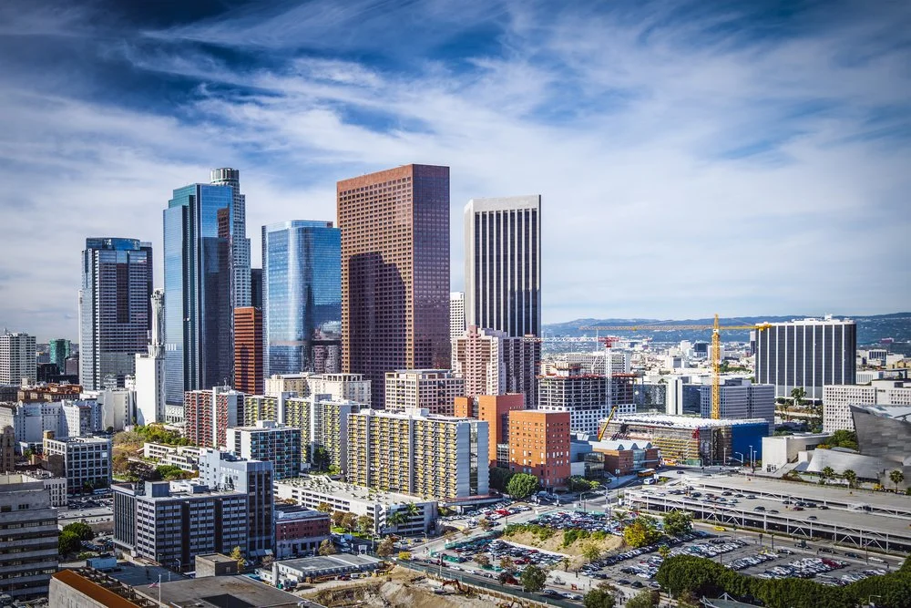 A sunny daytime cityscape featuring tall modern skyscrapers and construction cranes in downtown Los Angeles with a partly cloudy sky.
