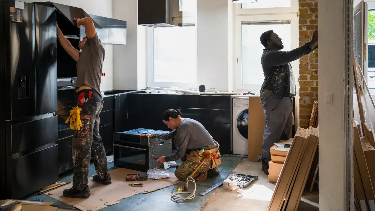 Three workers installing kitchen appliances and cabinetry in a home under construction, with one worker installing a refrigerator, another working on an oven, and the third assembling kitchen cabinets.