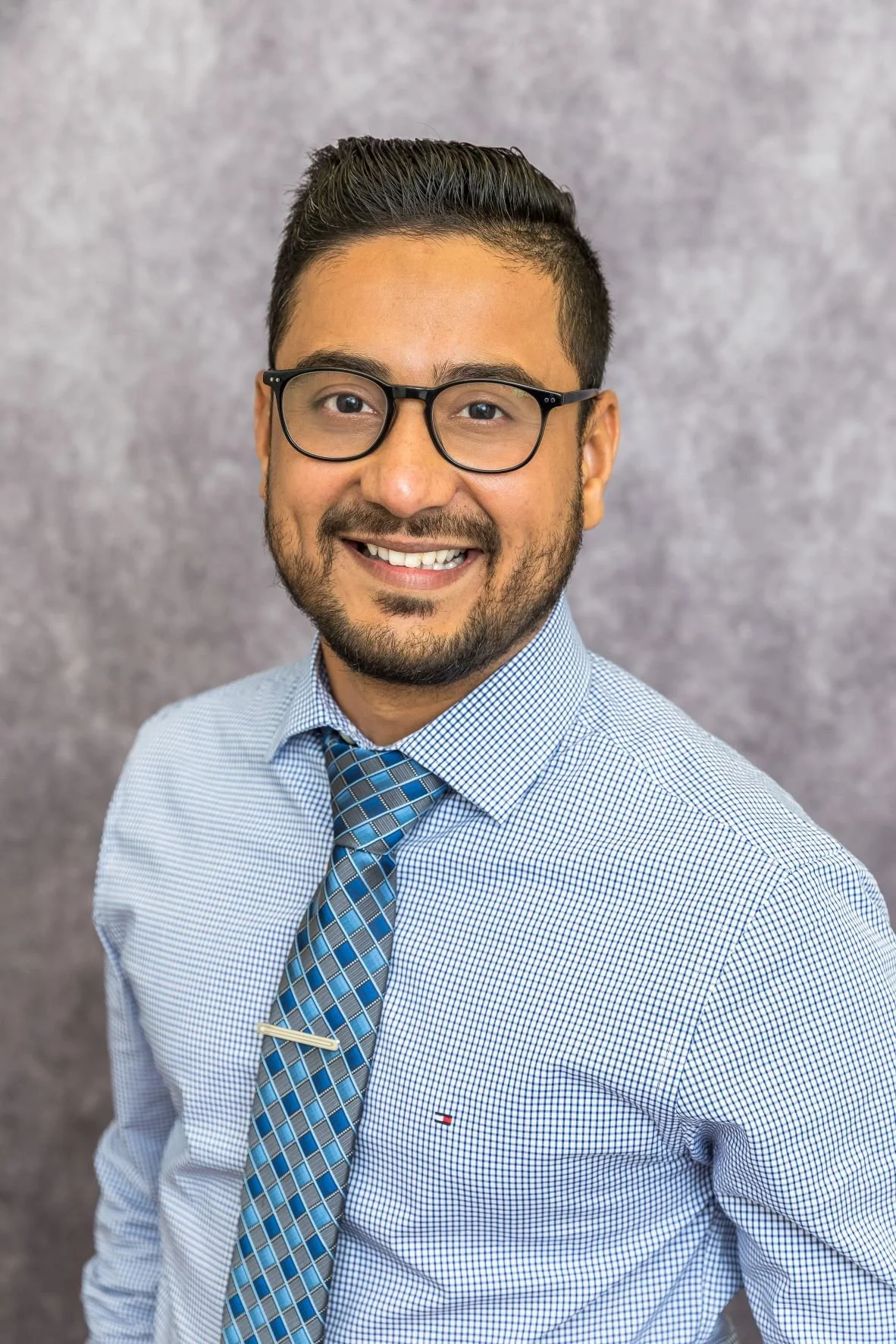 A smiling man with glasses, wearing a blue checkered shirt and a patterned tie, standing against a plain gray background.