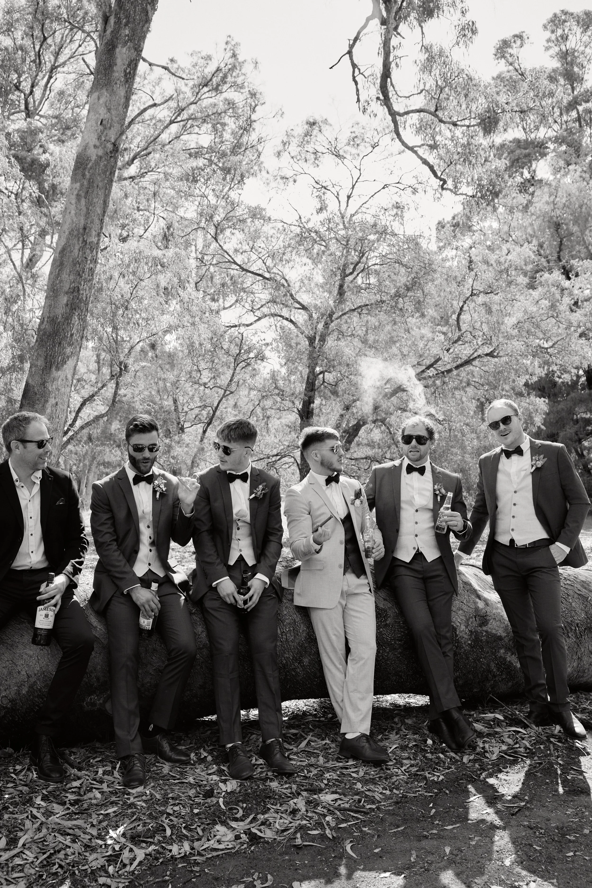 A group of seven men in suits and sunglasses, sitting and standing on a fallen log outdoors among trees, having drinks and chatting.