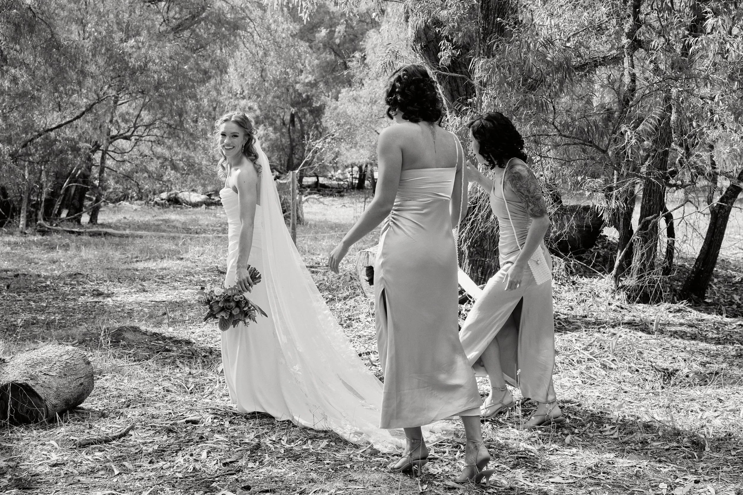 A black and white photograph of a bride in a wedding dress holding a bouquet and smiling, with three women in casual dresses and high heels, standing outdoors near trees and fallen logs.