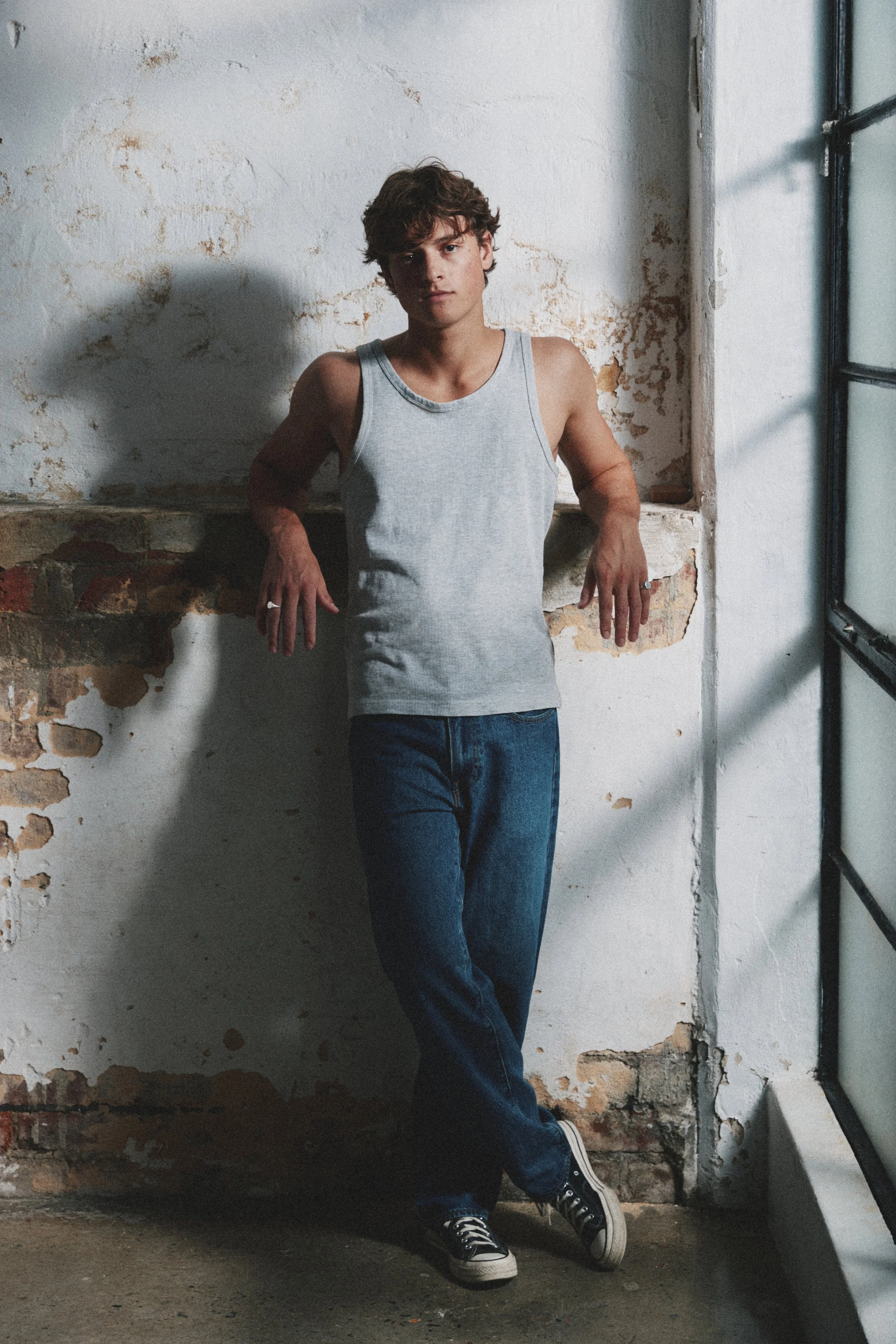 A young man with curly hair leaning against a textured, partially exposed brick wall, wearing a gray tank top, jeans, and black and white sneakers, inside an industrial-style space.