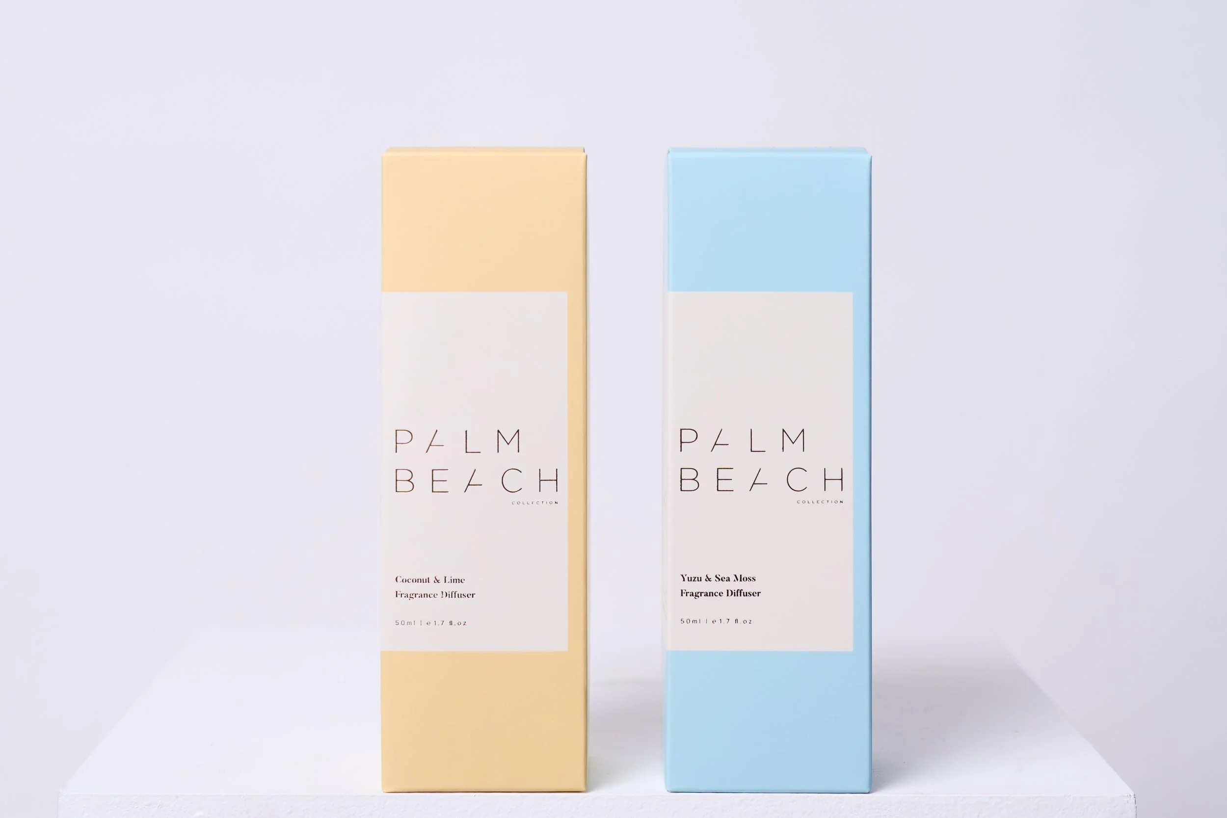 Two pastel-colored boxes of Palm Beach fragrance diffusers, one in pale yellow with coconut and lime scent, and one in pale blue with yuzu and sea moss scent, standing on a white shelf against a plain white background.