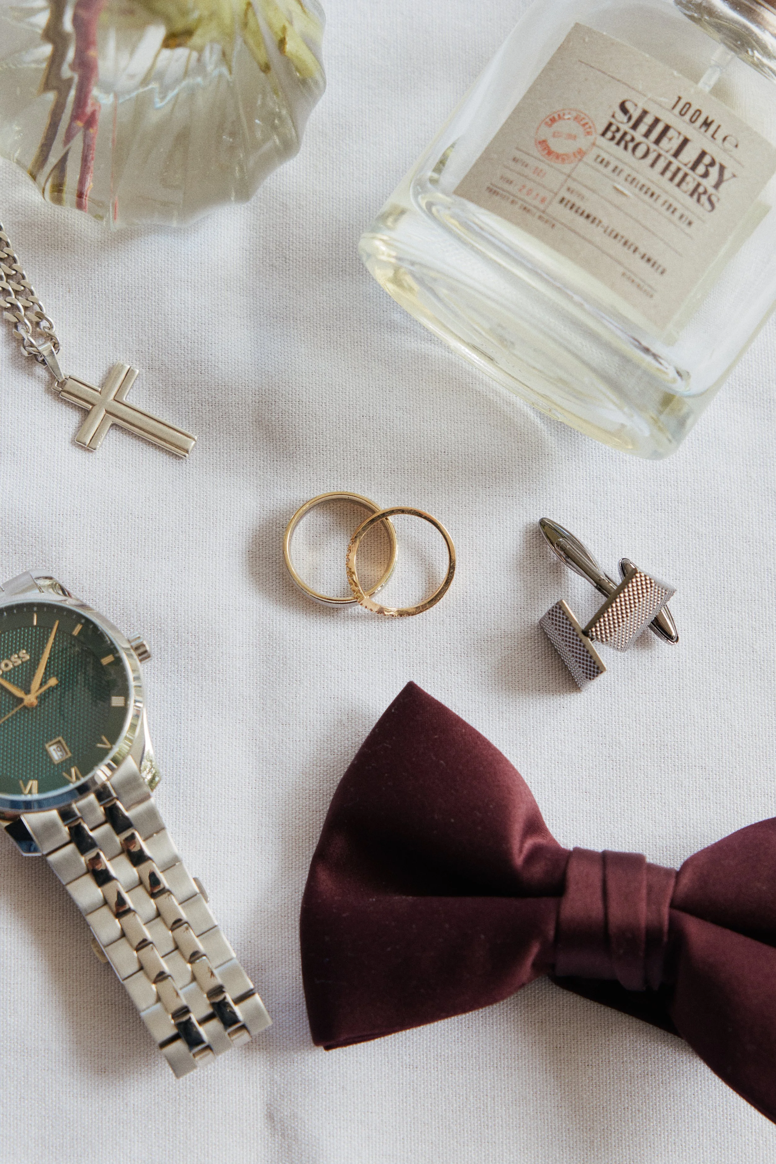 A collection of men's accessories and personal items including a silver wristwatch, two gold rings, a pair of silver cufflinks, a gold chain with a cross pendant, a burgundy bow tie, and a bottle of Shelby Brothers whiskey on a white surface.