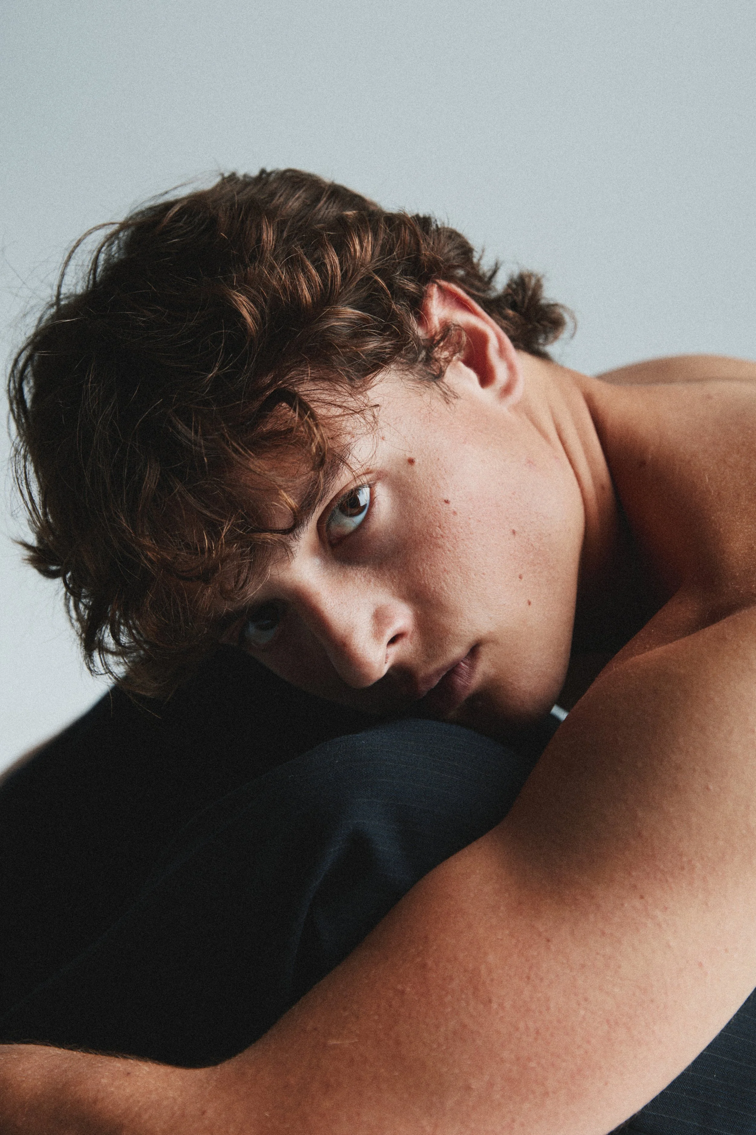 Close-up of a person's face with brown curly hair, blue eyes, and light skin, lying down on their side against a plain light background.