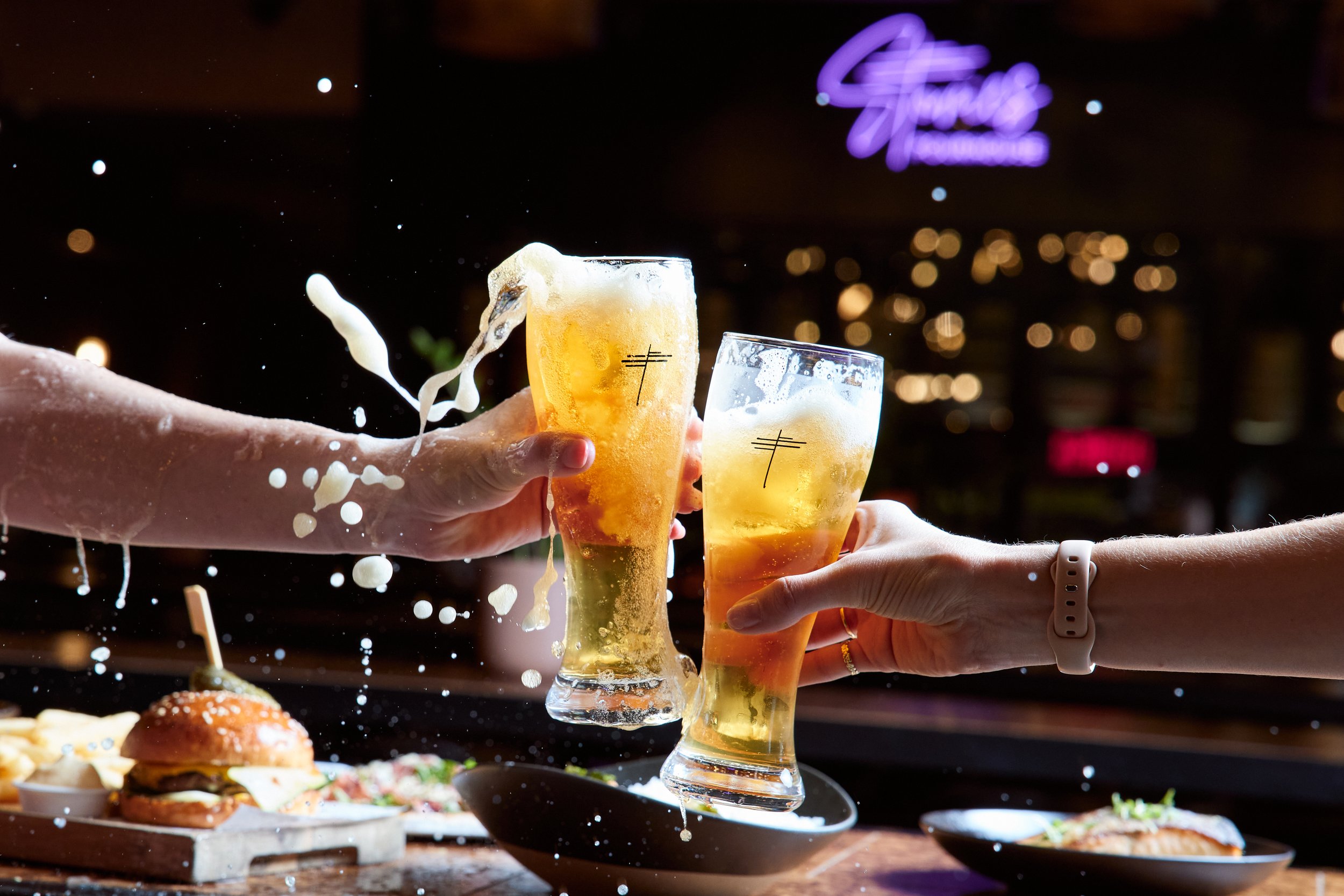 Two people clinking glasses of beer while spilling foam and liquid, with food on the table in the background, in a lively bar or restaurant setting.