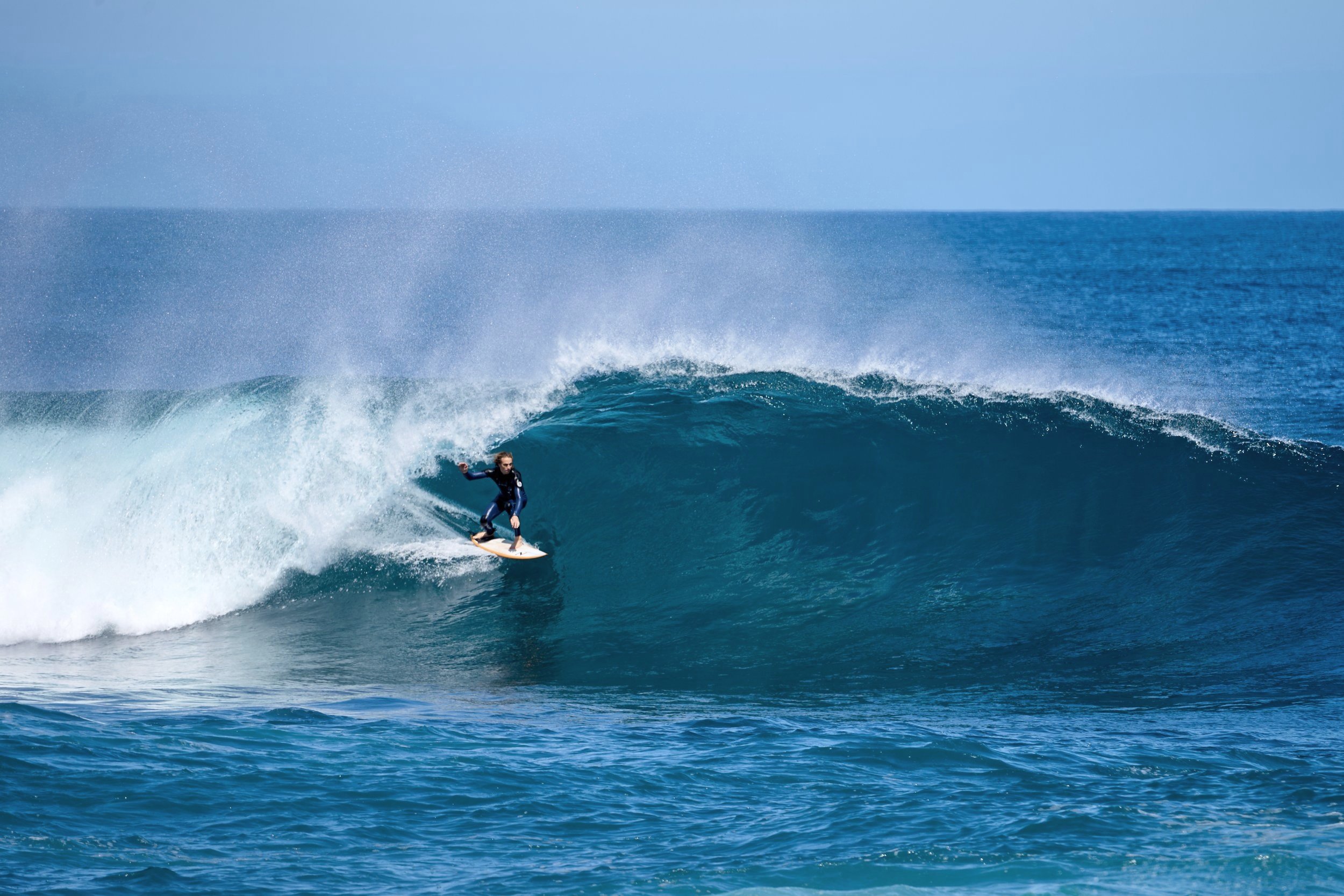 A person surfing on a large blue wave in the ocean under a clear sky.
