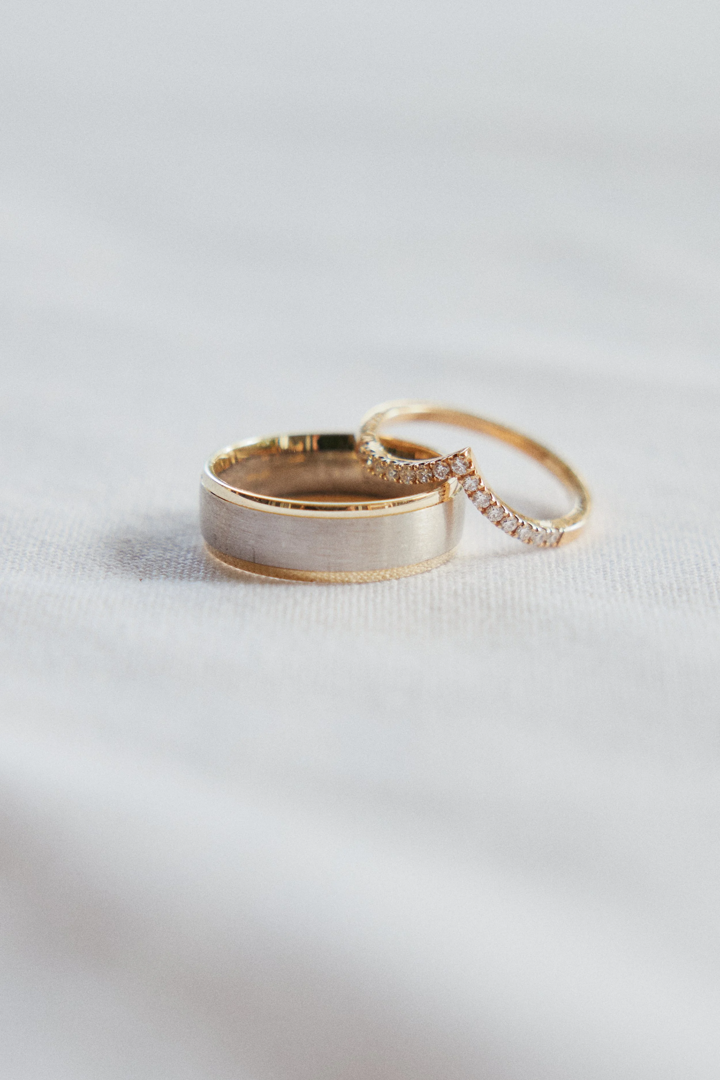 A wedding band with a silver center and gold edges and an engagement ring with small diamonds on a white fabric background.