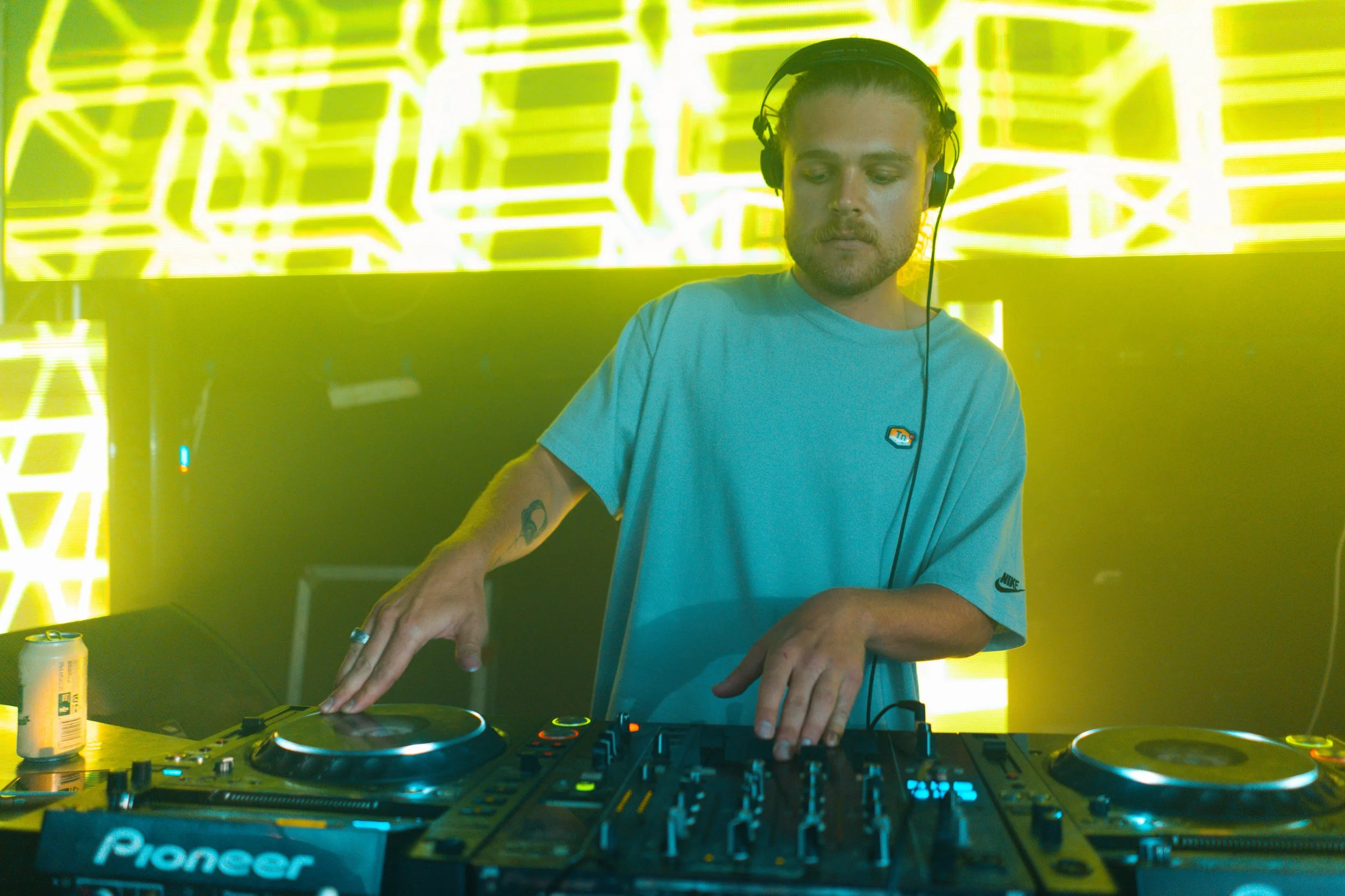 A male DJ wearing headphones and a light blue t-shirt is operating a Pioneer DJ controller on a table in front of a bright yellow neon-lit backdrop.
