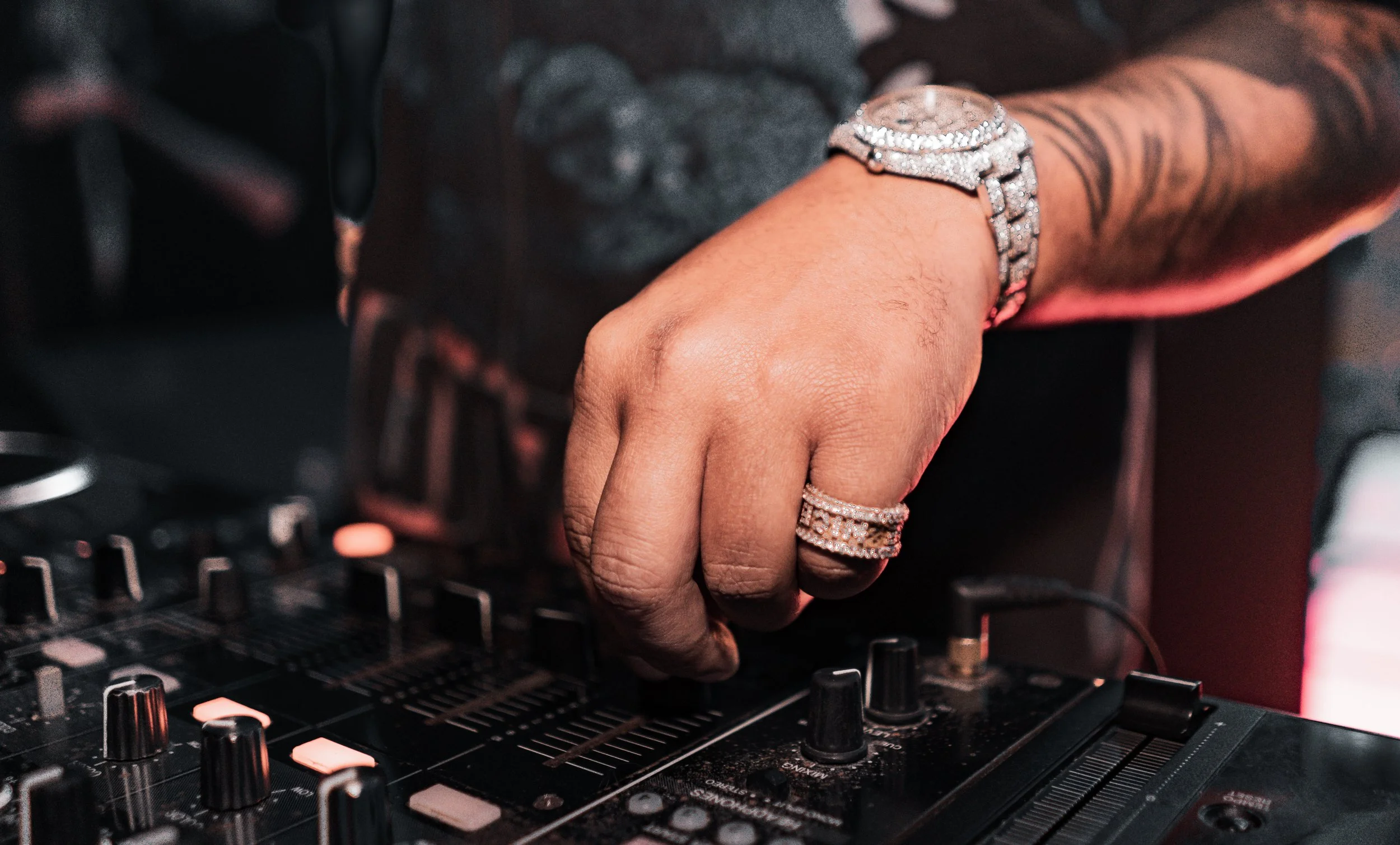 Close-up of a DJ's hand adjusting controls on a DJ mixer, wearing a watch and rings, with tattoos visible on the arm.