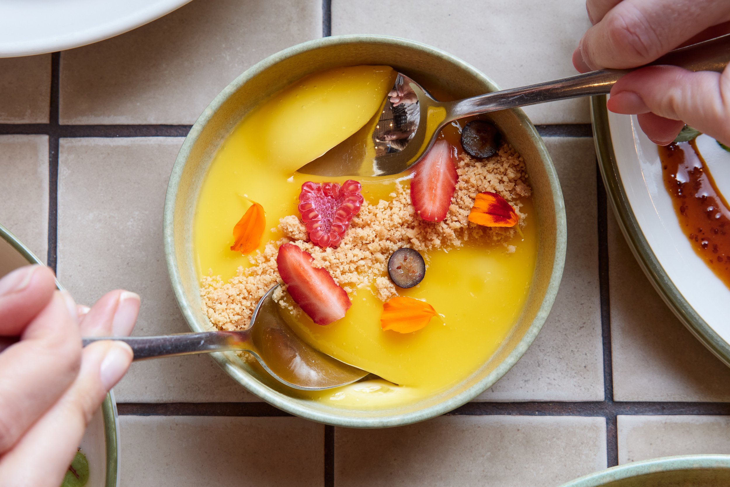 A bowl of yellow custard with fresh strawberries, raspberries, blueberries, edible flower petals, and crushed cookies or graham crackers.
