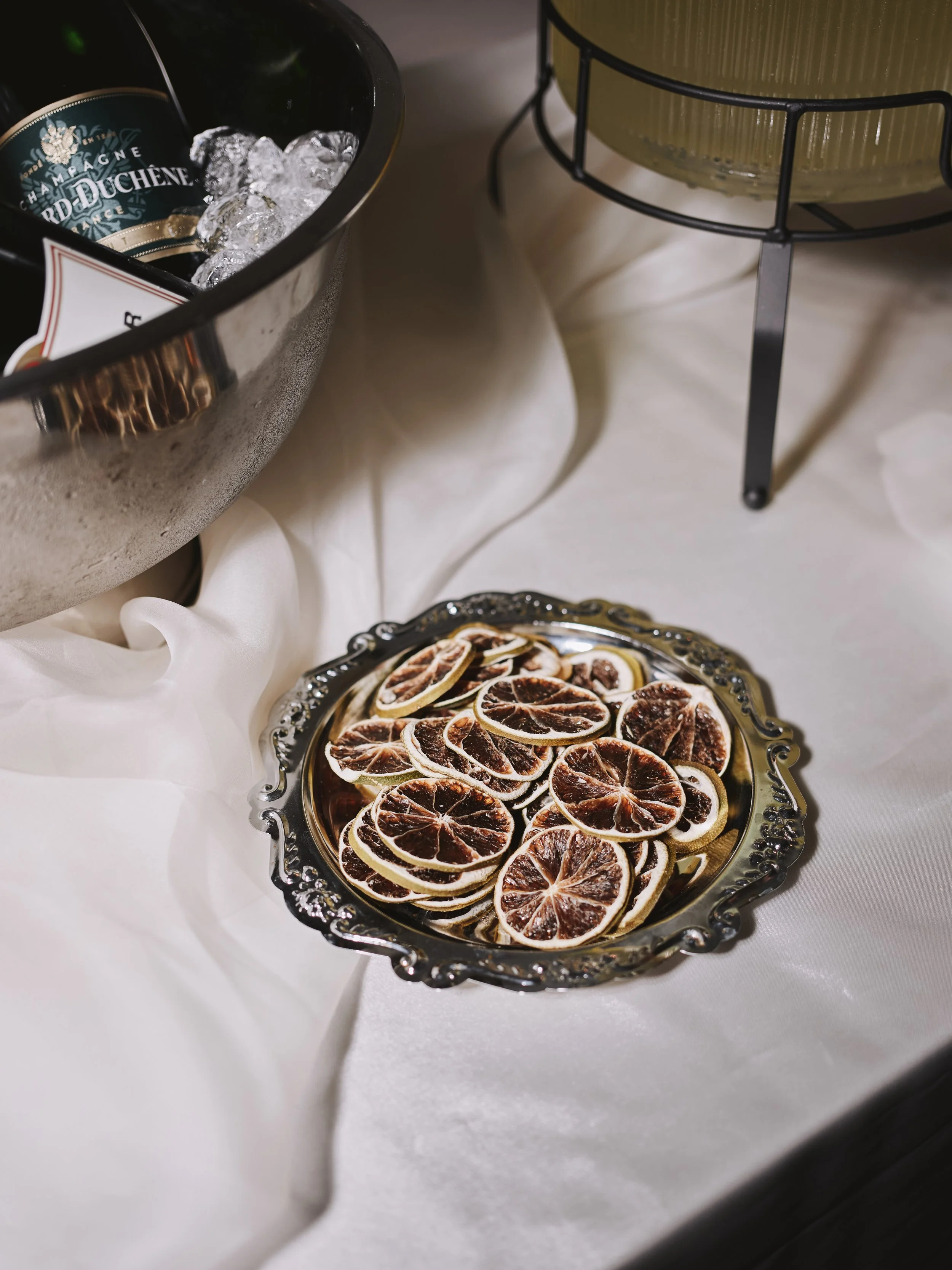 A silver tray filled with dried blood orange slices on a white tablecloth, with a metal bucket of ice and a champagne bottle in the background.