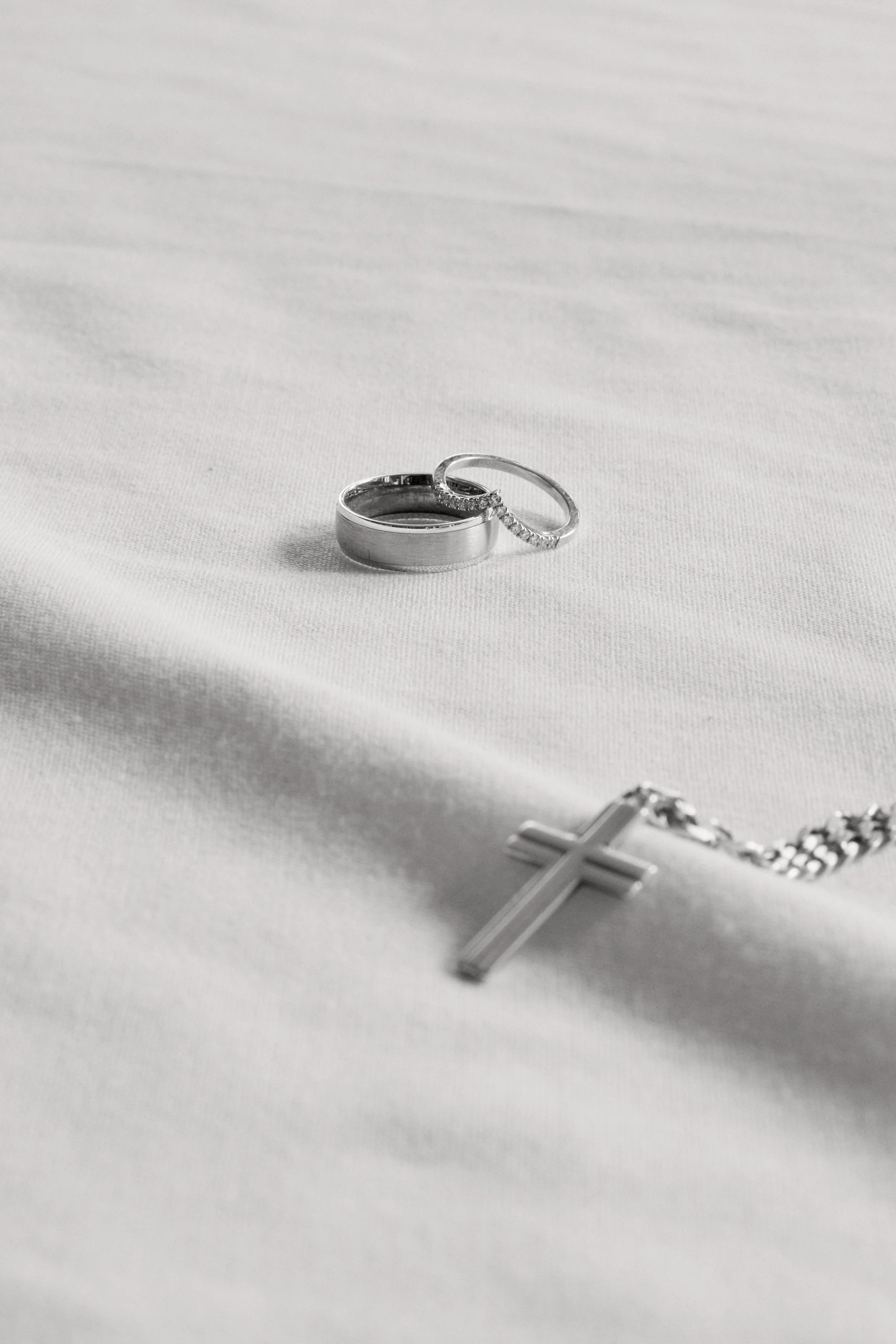 Silver wedding rings and a silver cross necklace resting on a white cloth background.