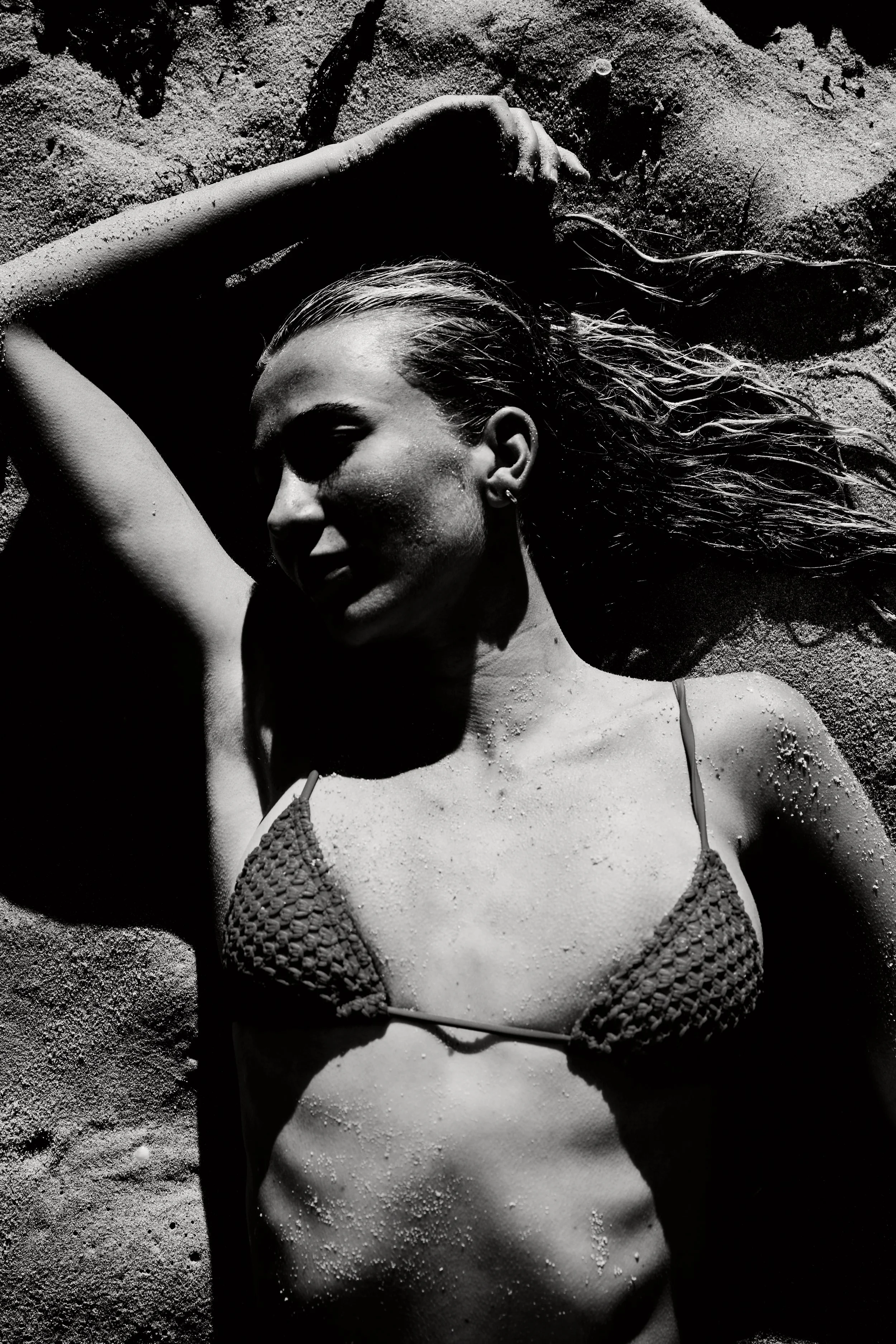 A woman lying on the sand at the beach, wearing a bikini, with her eyes closed and her hair wet, surrounded by sand grains.