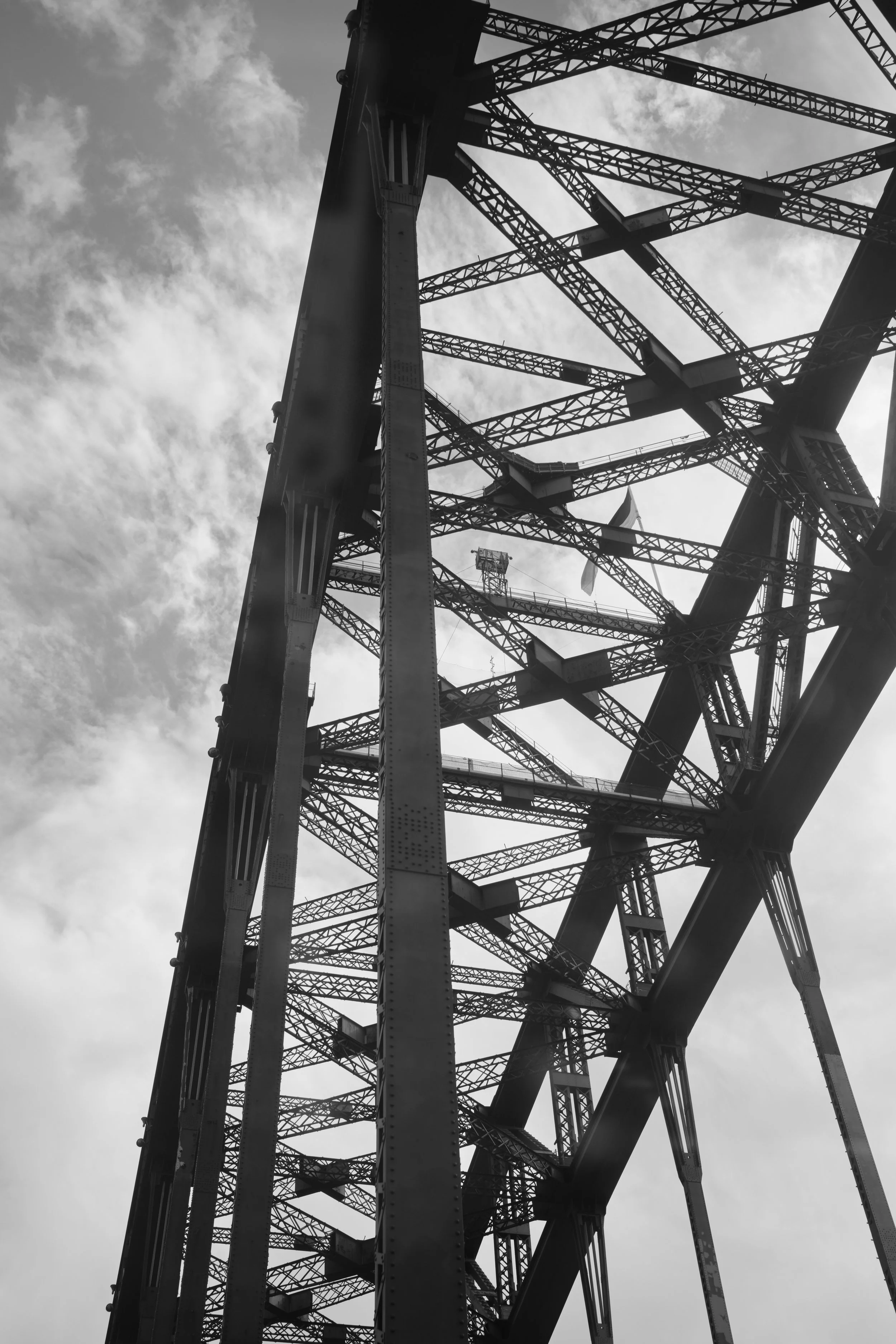 A black and white photo of the sydney harbour bridge from below, showcasing its metal structure and intricate framework against a cloudy sky.